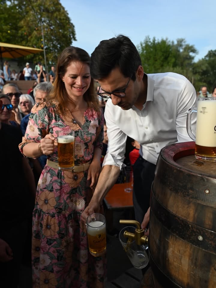 Zwei Personen stehen im Freien bei einem Fest. Eine Person in einem Blumenkleid hält ein volles Bierglas, während eine andere Person in weißem Hemd Bier aus einem großen Holzfass in ein Glas schenkt. Auf dem Fass steht ein weiteres volles Glas mit der Aufschrift „GANTER“. (KI-Alternativtext)