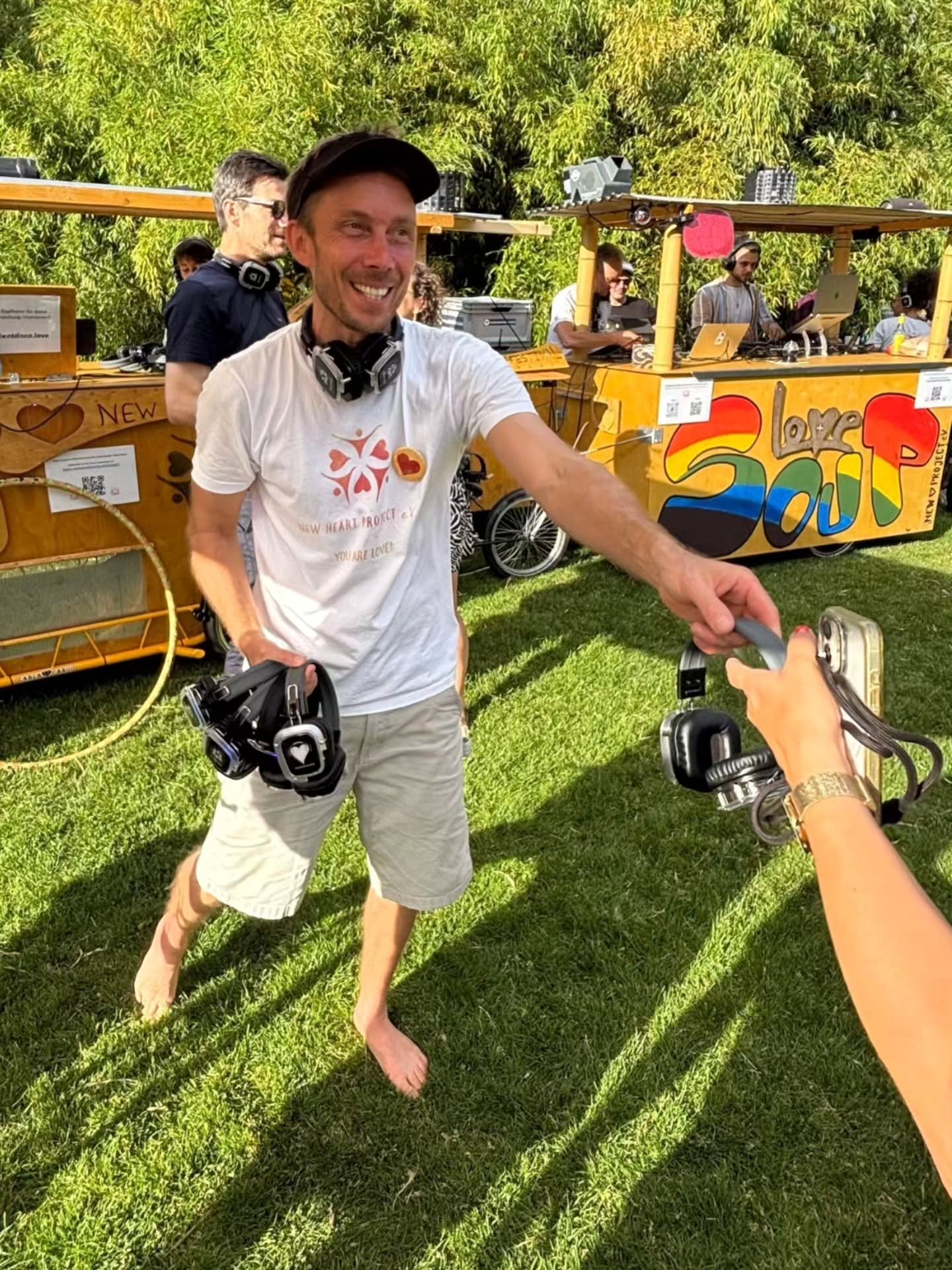 Ein lächelnder Mensch mit Kopfhörern um den Hals reicht einer anderen Person Kopfhörer, im Hintergrund ein farbenfroher DJ-Container mit der Aufschrift „Love Soup“ und weitere Personen auf einer Wiese. (KI-Alternativtext)