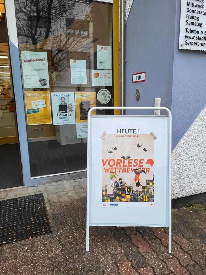 Dieser Alternativtext wurde KI-generiert: Ein Schaufenster mit Aushängen und ein A-Ständer davor. Der A-Ständer bewirbt eine Vorleseaktion mit einem bunten Bild.