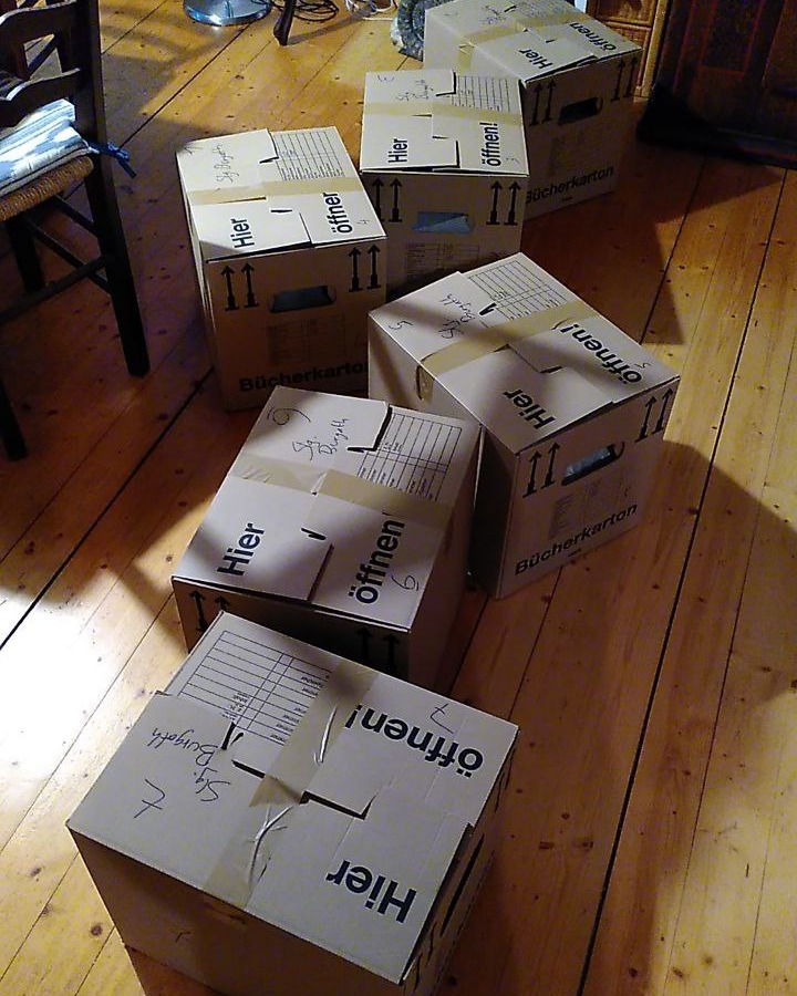 Dieser Alternativtext wurde KI-generiert: Mehrere braune Kartons mit Pfeilen und Text sind auf einem Holzboden gestapelt. Die Kartons sind mit dem Wort "Hier" und Anweisungen zum Öffnen beschriftet.