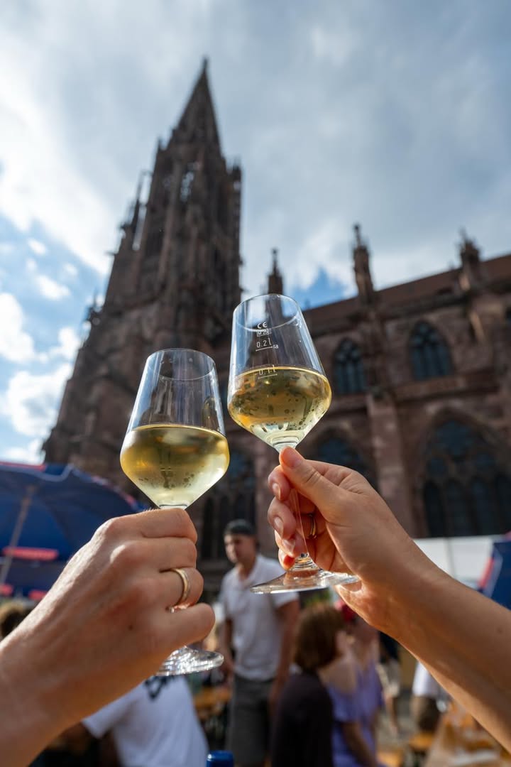 Zwei Hände halten Gläser mit Weißwein und stoßen im Vordergrund an. Im unscharfen Hintergrund erhebt sich eine große gotische Kirche über einer Menschenmenge. (KI-Alternativtext)