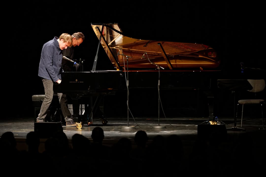 Dieser Alternativtext wurde KI-generiert: Schwarz-Weiß-Fotografie einer Bühnenaufführung. Eine Person spielt Klavier, während im Hintergrund ein Publikum sitzt.