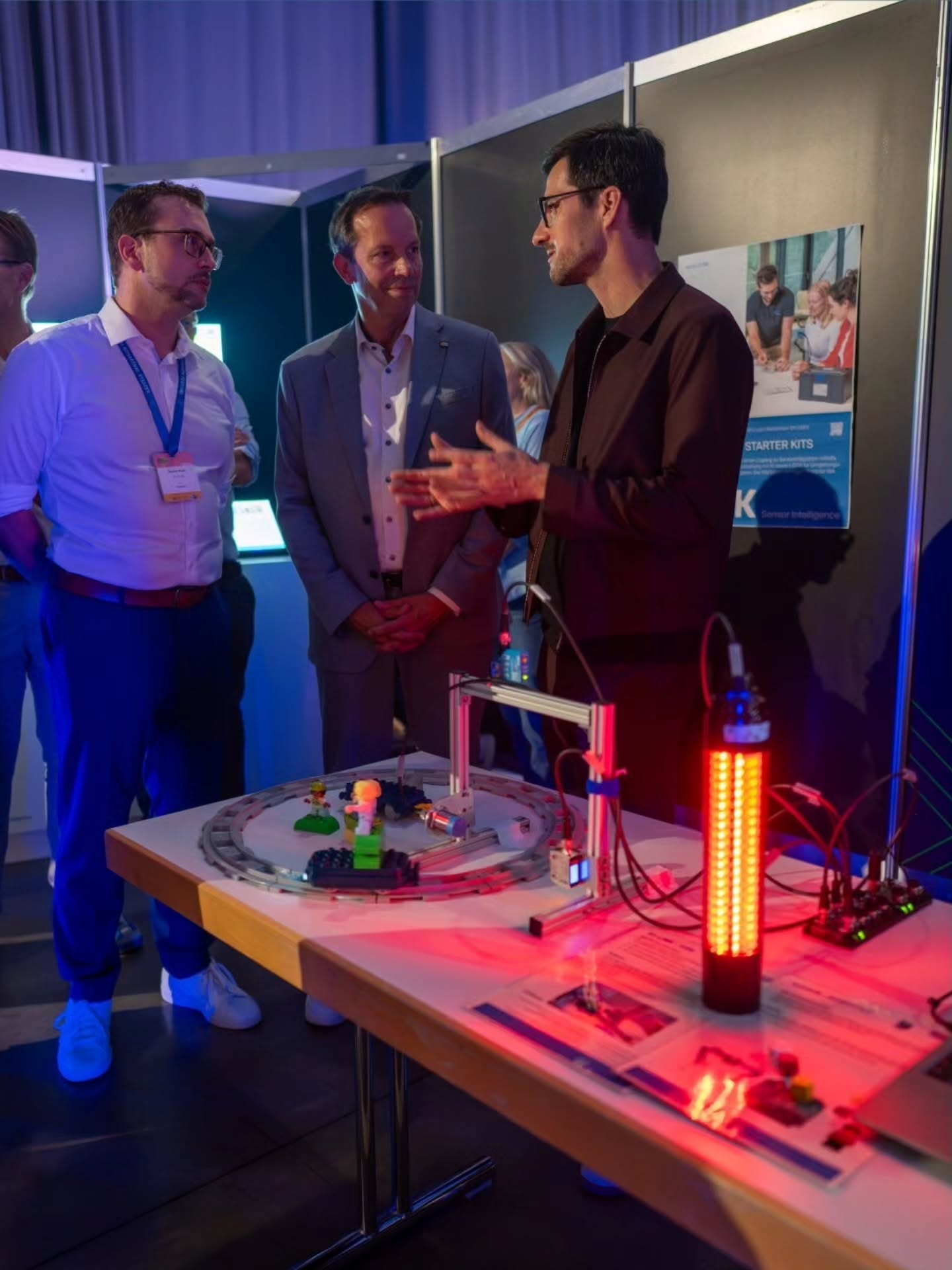 Drei Personen stehen an einem Tisch auf einer Messe und betrachten eine technische Demonstration. Eine Person erklärt mit Gesten die Funktion der Ausstellungsstücke, zu denen eine Modellbahn mit Figuren, ein Metallgestell und ein rot leuchtender Sensorzylinder gehören. Im Hintergrund ist ein Plakat mit dem Text "STARTER KITS" und "Sensor Intelligence" sichtbar. (KI-Alternativtext)