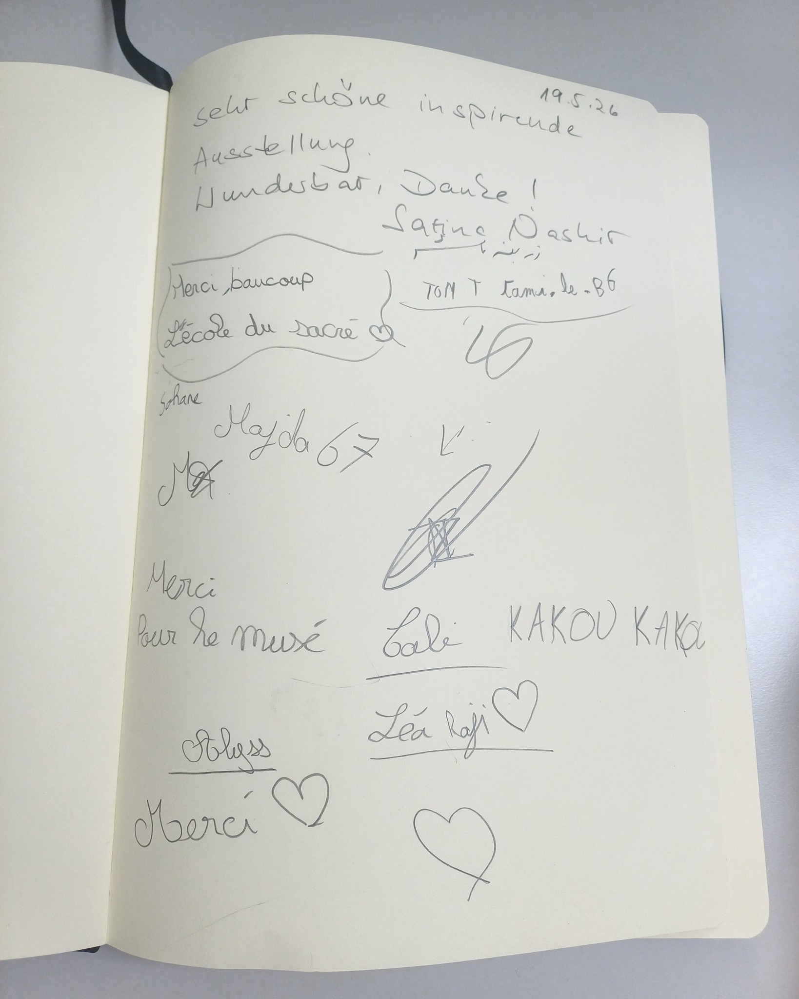 Eine Seite eines offenen Notizblocks zeigt zahlreiche handschriftliche Einträge, Unterschriften und Dankesworte. Oben steht das Datum 19.5.26 und der Text "sehr schöne inspirierende Ausstellung. Wunderbar, Danke!". Weitere Einträge enthalten Grußformeln wie "Merci beaucoup" und "Merci Pour le musé" sowie Namen wie Latina Oashir, Sohane, Hayda 67, KAKOU KAKA und Lea Raji, teilweise ergänzt durch gezeichnete Herzen. (KI-Alternativtext)