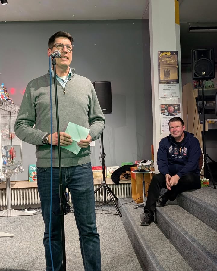 Dieser Alternativtext wurde KI-generiert: Eine Person hält ein Mikrofon und spricht, während eine andere Person im Hintergrund sitzt. Im Hintergrund sind Dekorationen und Geräte zu sehen.