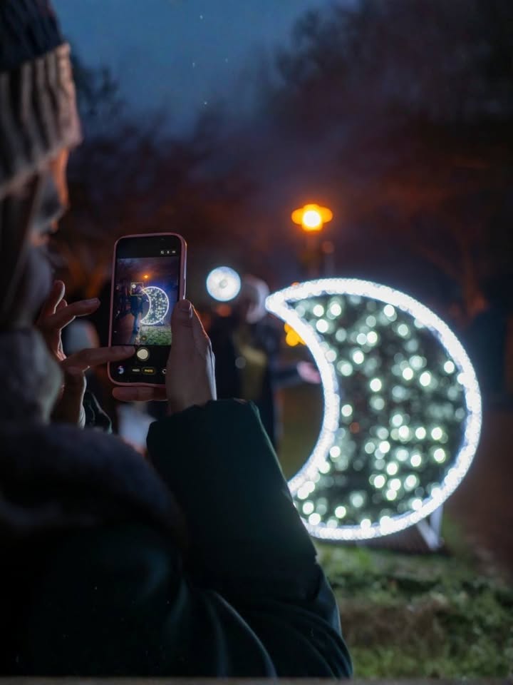 Dieser Alternativtext wurde KI-generiert: Eine Person hält ein Smartphone hoch und fotografiert eine leuchtende Halbmond-Installation. Im Hintergrund sind weitere Lichtquellen und Bäume erkennbar.