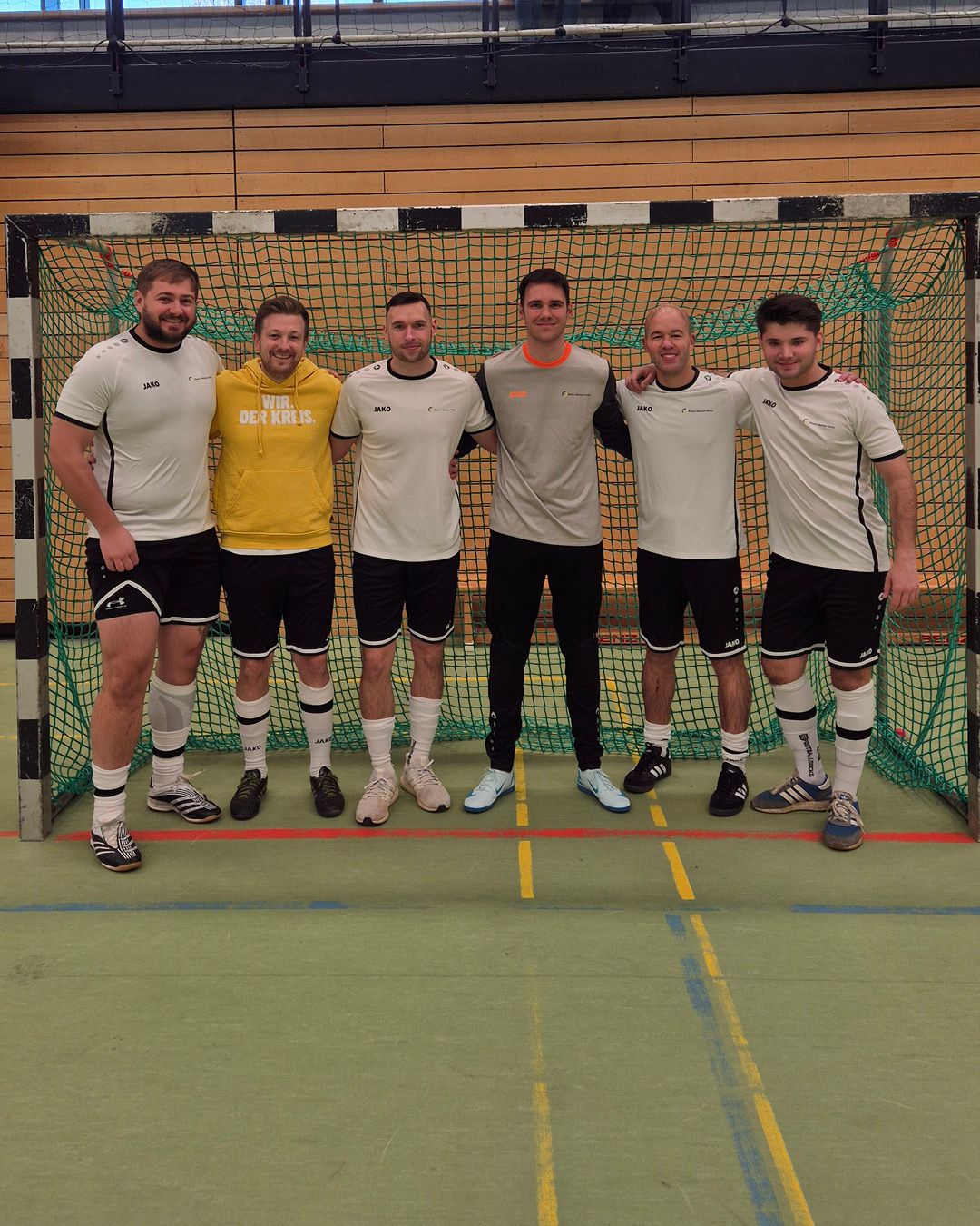 Dieser Alternativtext wurde KI-generiert: Eine Gruppe von Personen in Sportkleidung steht nebeneinander in einer Halle. Der Hintergrund besteht aus einer Wand mit bunten Feldern und einem Tor.