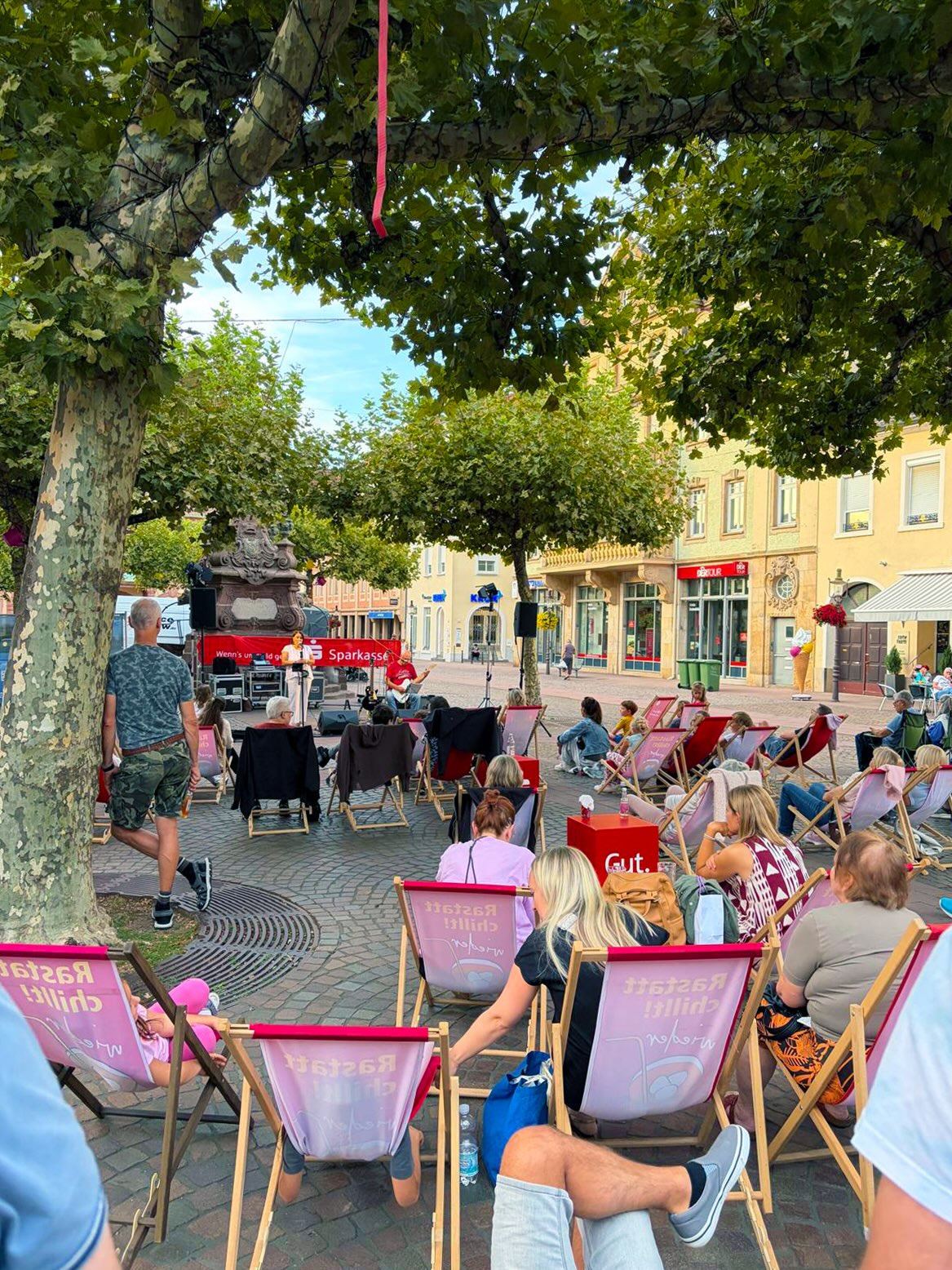 Personen sitzen auf einem gepflasterten Platz in Liegestühlen mit der Aufschrift „Raststatt chillt!“ und schauen auf eine Bühne, wo eine Person spricht und eine andere Gitarre spielt. Im Hintergrund ist ein rotes Banner der Sparkasse zu sehen. (KI-Alternativtext)