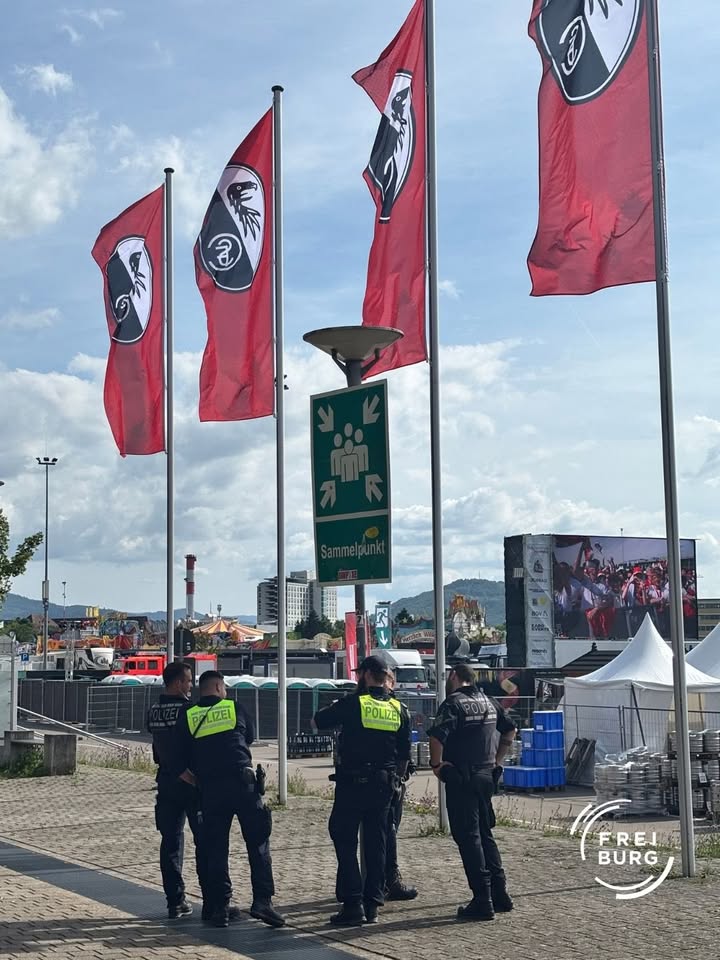 Mehrere Polizisten in Uniform stehen vor roten Fahnen mit einem schwarzen Vogel-Logo. Ein grünes Schild weist auf einen Sammelpunkt hin, im Hintergrund sind Zelte, ein Bildschirm und eine Stadtlandschaft zu sehen. (KI-Alternativtext)