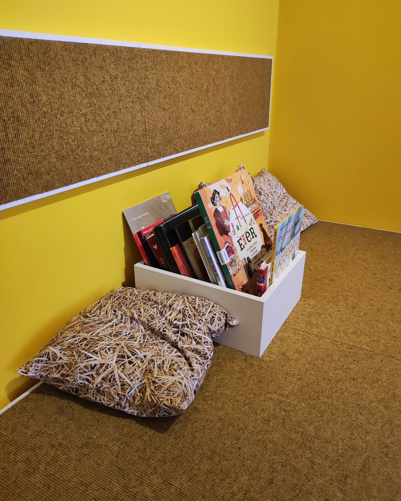 Dieser Alternativtext wurde KI-generiert: Ein Stapel Bücher steht in einer weißen Kiste, umgeben von Kissen mit Strohfüllung. Der Raum hat gelbe Wände und einen hellbraunen Boden.
