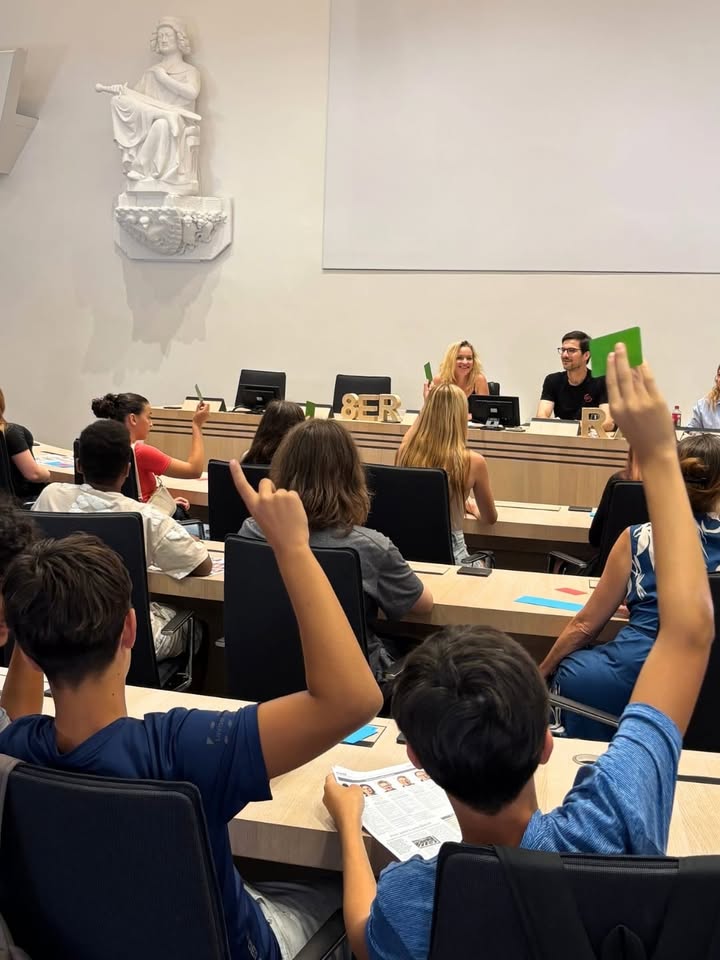 In einem Klassenzimmer sitzen mehrere Personen an Tischen, wobei zwei Personen im Vordergrund die Hände heben und eine eine grüne Karte hält. An der Vorderseite sitzen zwei weitere Personen hinter einem Tisch mit Computern und den Buchstaben BER, wobei eine Person ebenfalls eine grüne Karte hochhält. An der linken Wand ist eine weiße Statue angebracht. (KI-Alternativtext)