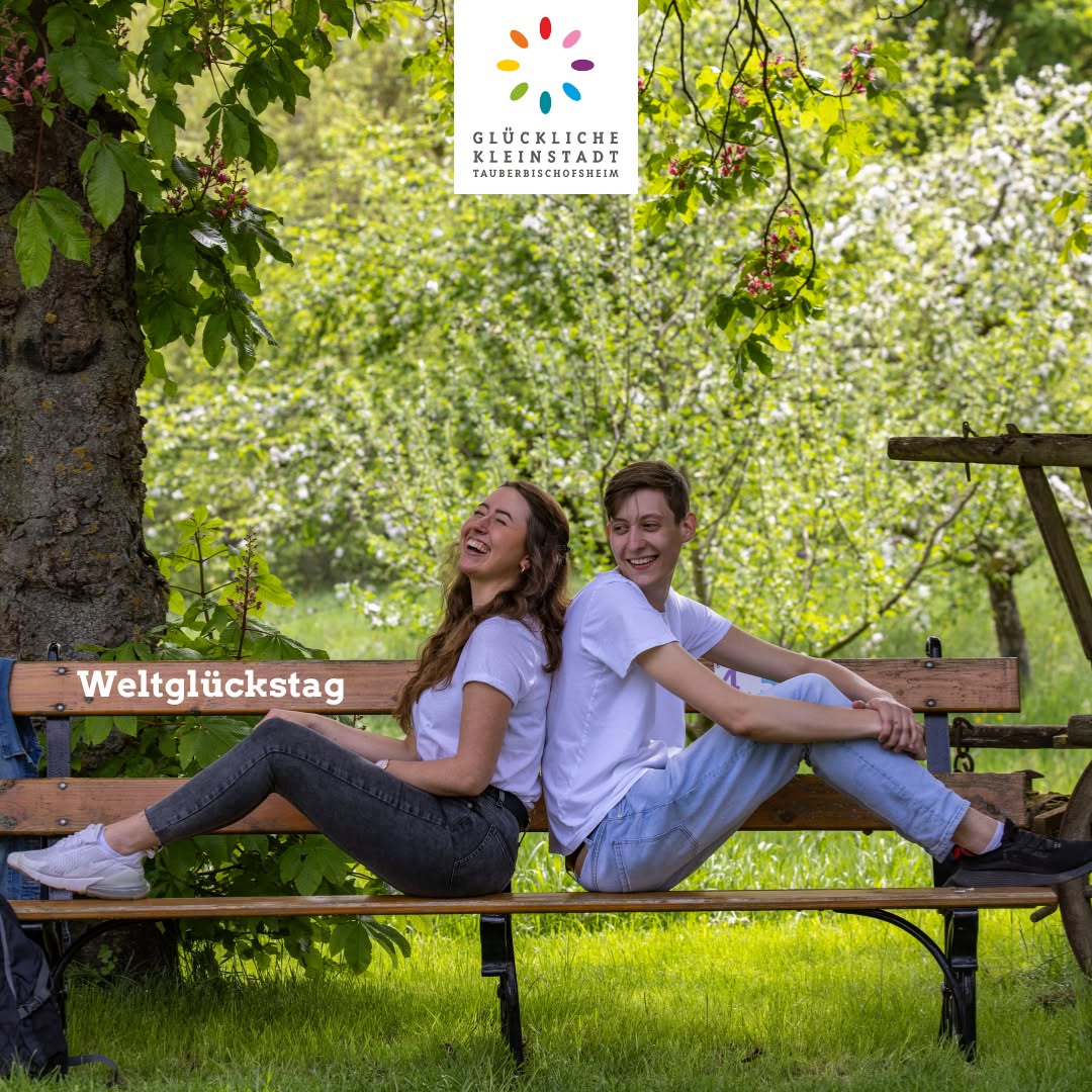 Dieser Alternativtext wurde KI-generiert: Zwei Personen sitzen lächelnd auf einer Parkbank unter Bäumen und Grün. Das Bild wirkt fröhlich und entspannt.