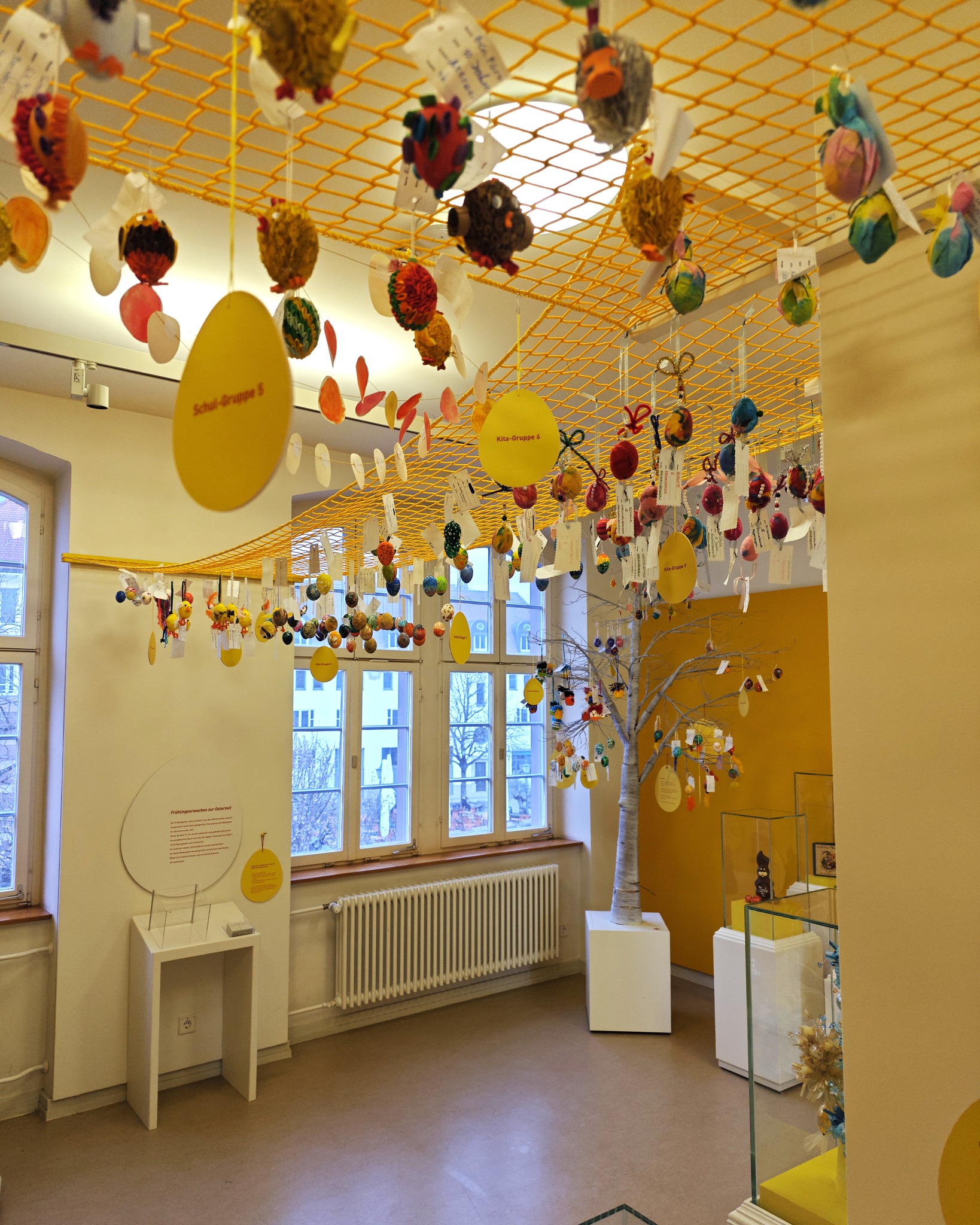 Dieser Alternativtext wurde KI-generiert: Ein heller Raum mit einer gelben Decke, von der viele bunte, stoffähnliche Objekte hängen. Im Raum stehen Bäume und Schilder mit Text.