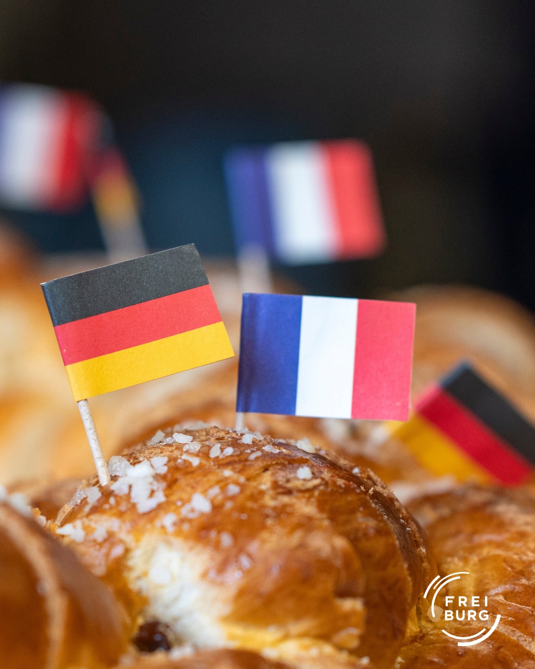 Dieser Alternativtext wurde KI-generiert: Mehrere kleine Flaggen stehen in einem Gebäckstück. Im Hintergrund sind weitere Flaggen unscharf erkennbar.