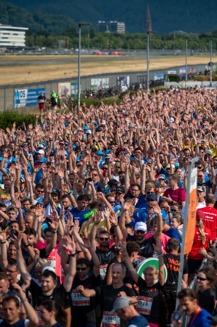 Eine große Menschenmenge von Läufern bei einem Massensportevent hebt die Hände in die Luft. Viele Teilnehmer tragen blaue Trikots und Startnummern, wie die sichtbaren Nummern 1960 und 1918. Im Hintergrund erstrecken sich eine Absperrung, Gebäude und bewaldete Hügel. (KI-Alternativtext)
