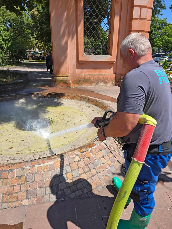 Eine Person mit grauem Haar und blauer Arbeitskleidung reinigt mit einem Hochdruckreiniger einen runden, gepflasterten Brunnen. Im Hintergrund steht ein orangefarbener Pavillon mit Gitterfenstern, dahinter sind Bäume und ein Gehweg zu sehen. (KI-Alternativtext)