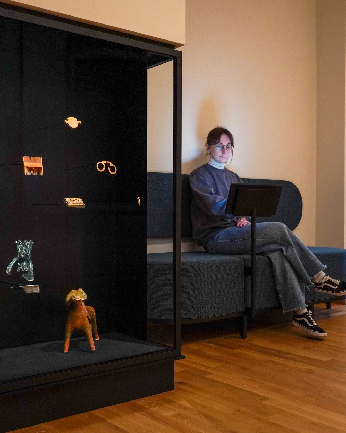 Dieser Alternativtext wurde KI-generiert: Eine Person sitzt mit einem Laptop auf einer Bank vor einer dunklen Vitrine mit verschiedenen Objekten darin. Der Raum ist hell erleuchtet und hat einen Holzboden.