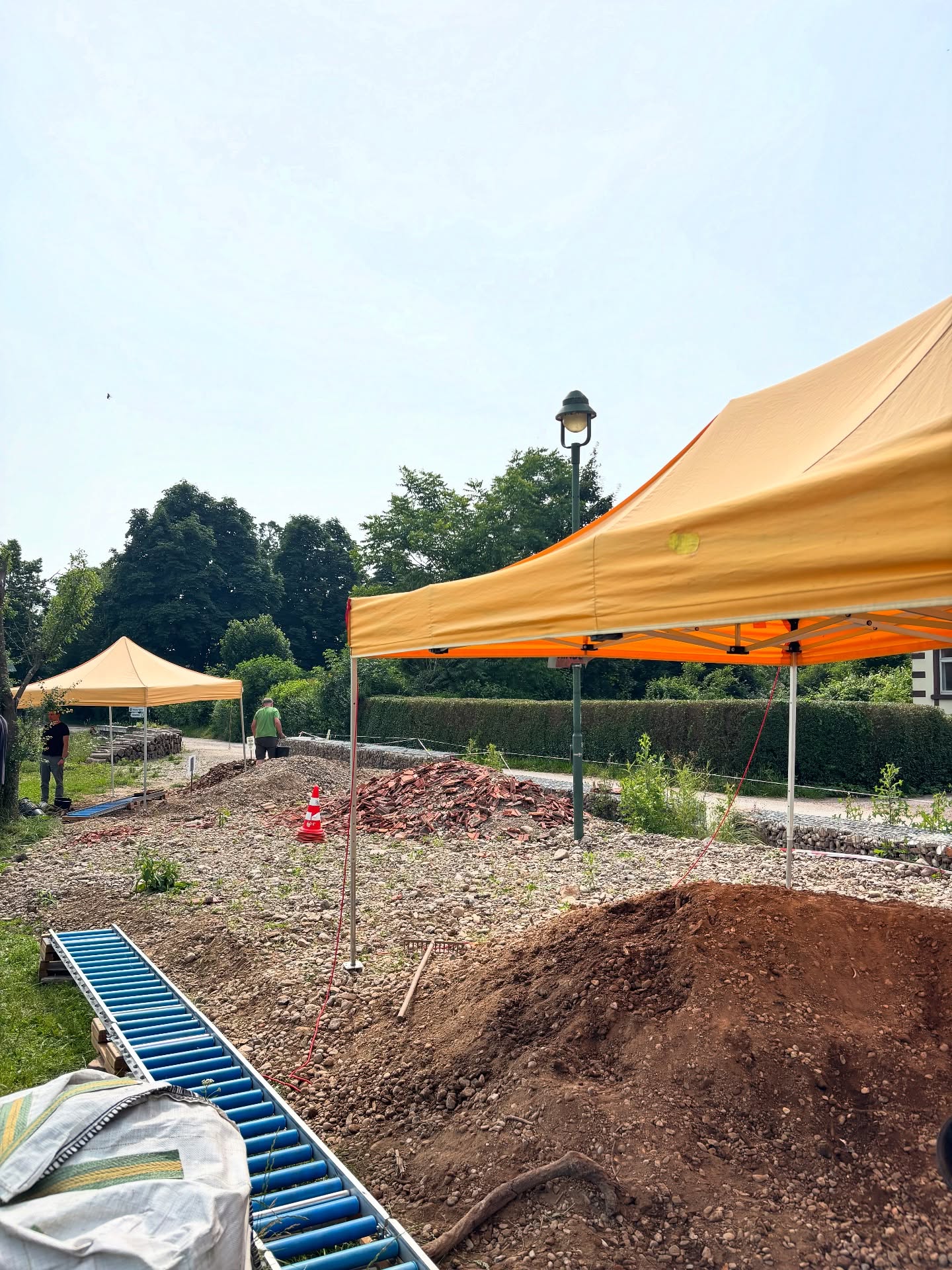Das Bild zeigt eine Baustelle im Freien mit zwei Pavillons, einem Haufen Erde und verschiedenen Werkzeugen. Es sind auch Bäume im Hintergrund zu sehen.