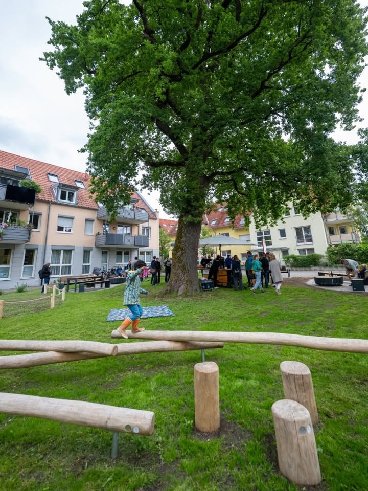 Ein Kind balanciert auf einem Holzstamm im Vordergrund eines Grünflächen-Spielplatzes, im Hintergrund stehen Menschen unter einem großen Baum und vor Wohnhäusern. (KI-Alternativtext)