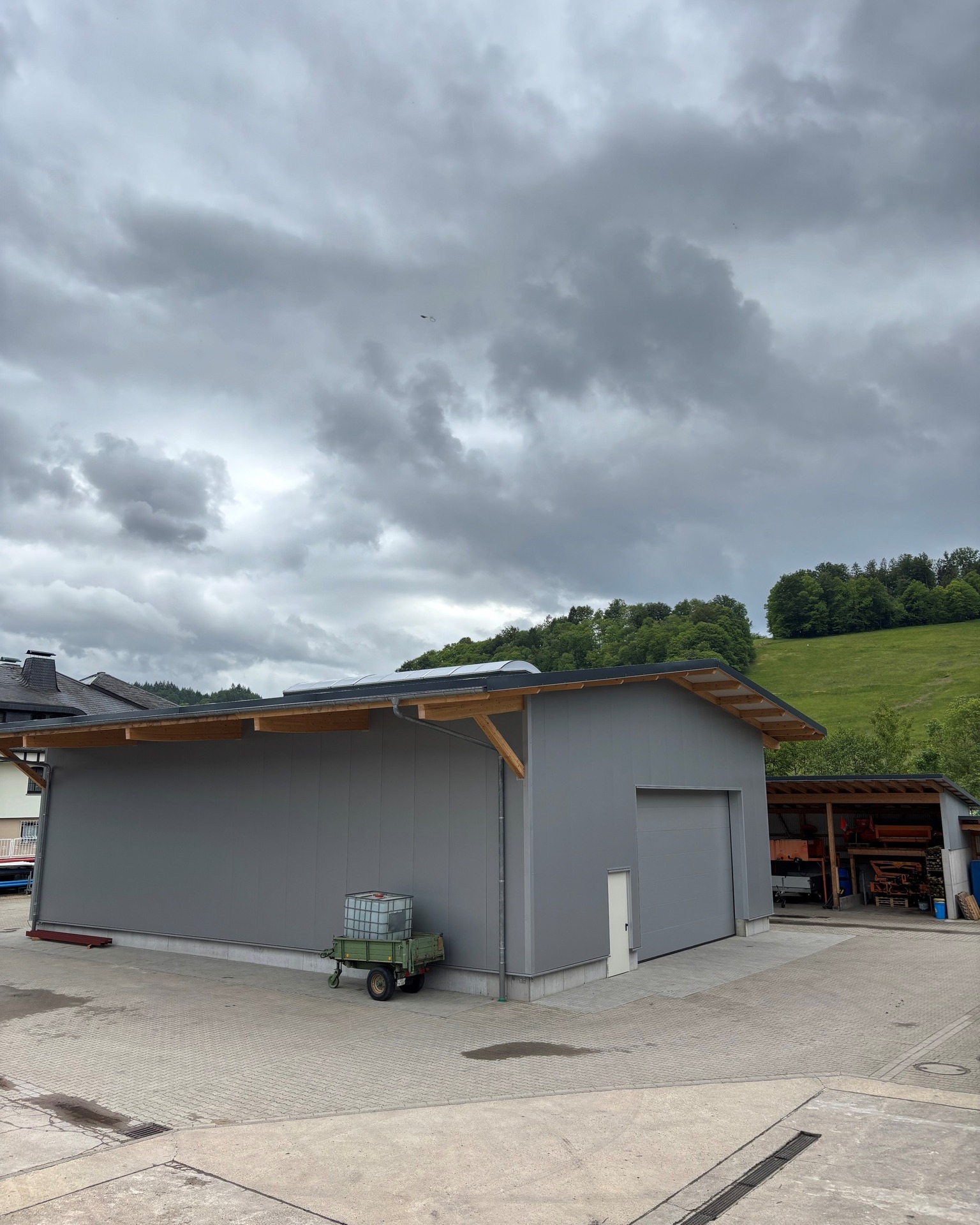 Graues Hallenbauwerk mit Holzvordach und Rolltor, daneben ein grüner Anhänger mit Behälter auf gepflastertem Hof. Im Hintergrund bewaldeter Hügel unter bedecktem Himmel. (KI-Alternativtext)