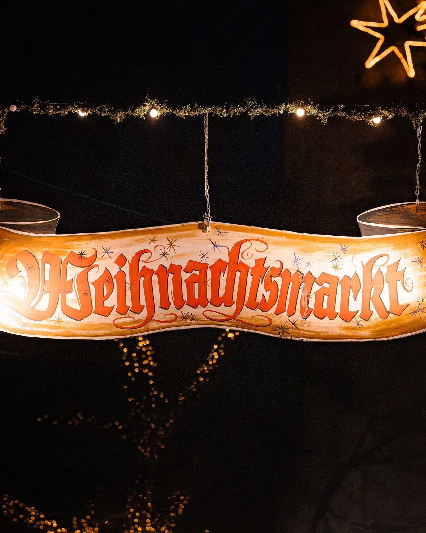 Ein beleuchtetes Schild mit der Aufschrift Weihnachtsmarkt hängt an Ketten. Es ist in einem geschwungenen Bannerdesign gestaltet.