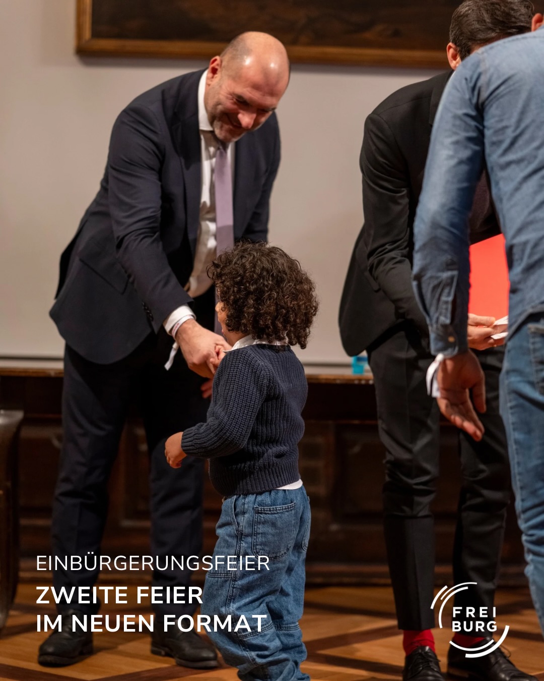 Dieser Alternativtext wurde KI-generiert: Ein farbiges Foto zeigt drei Personen in einem Innenraum. Eine Person kniet sich zu einem Kind, während eine andere im Hintergrund steht.