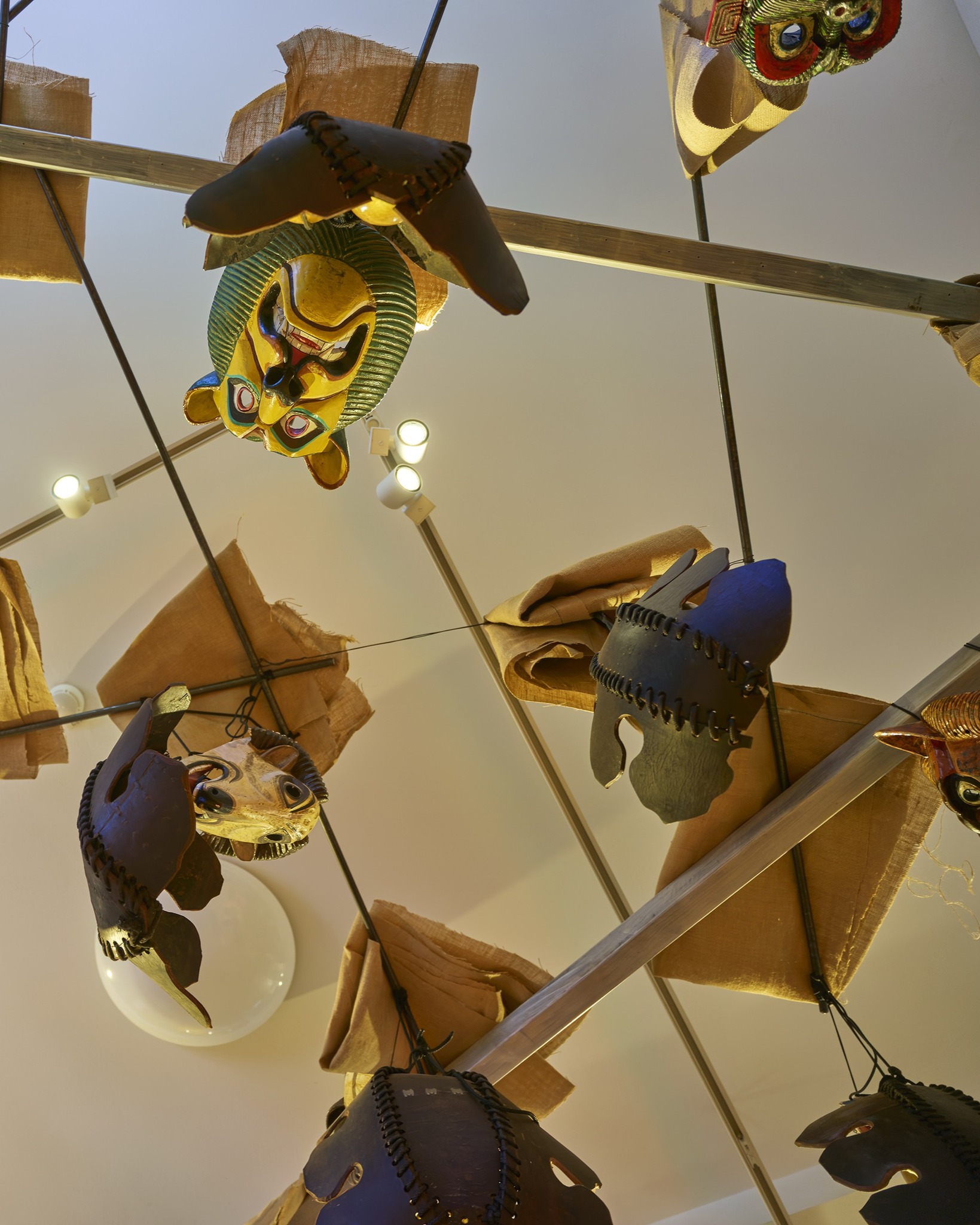 Mehrere Masken hängen an einem Metallgestell von der Decke herab. Dazu gehören eine goldene Maske mit grünem Kopfschmuck sowie dunkle Masken mit sichtbaren Nähten. Braune Stoffstücke und Deckenleuchten sind ebenfalls zu sehen. (KI-Alternativtext)