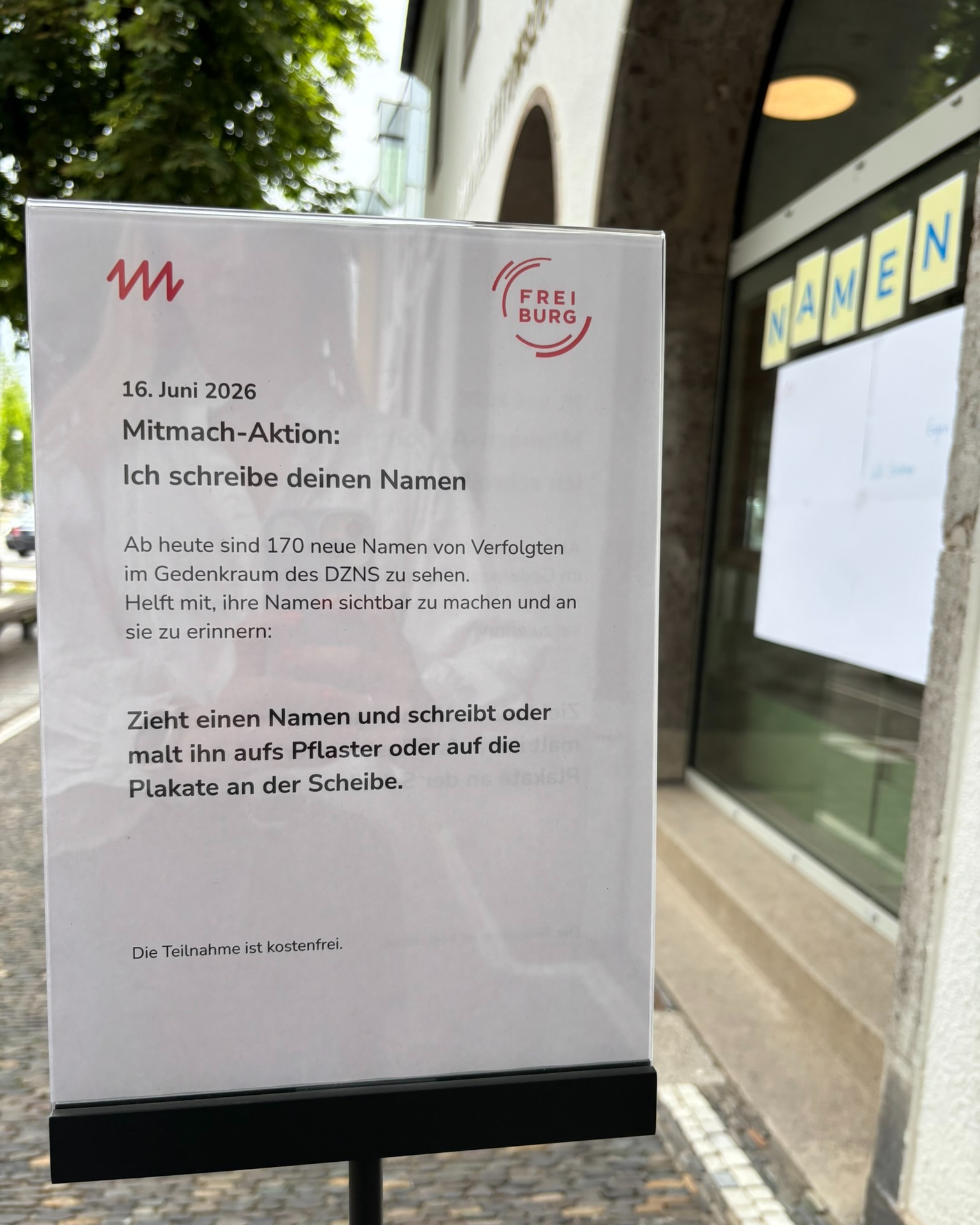 Ein weißes Informationsschild vor einem Schaufenster trägt die Überschrift „16. Juni 2026 Mitmach-Aktion: Ich schreibe deinen Namen“. Der Text informiert über 170 neue Namen von Verfolgten im Gedenkraum des DZNS und fordert zur Teilnahme auf, indem man diese Namen auf Pflastersteine oder Plakate schreibt. Am unteren Rand steht: „Die Teilnahme ist kostenfrei.“ (KI-Alternativtext)