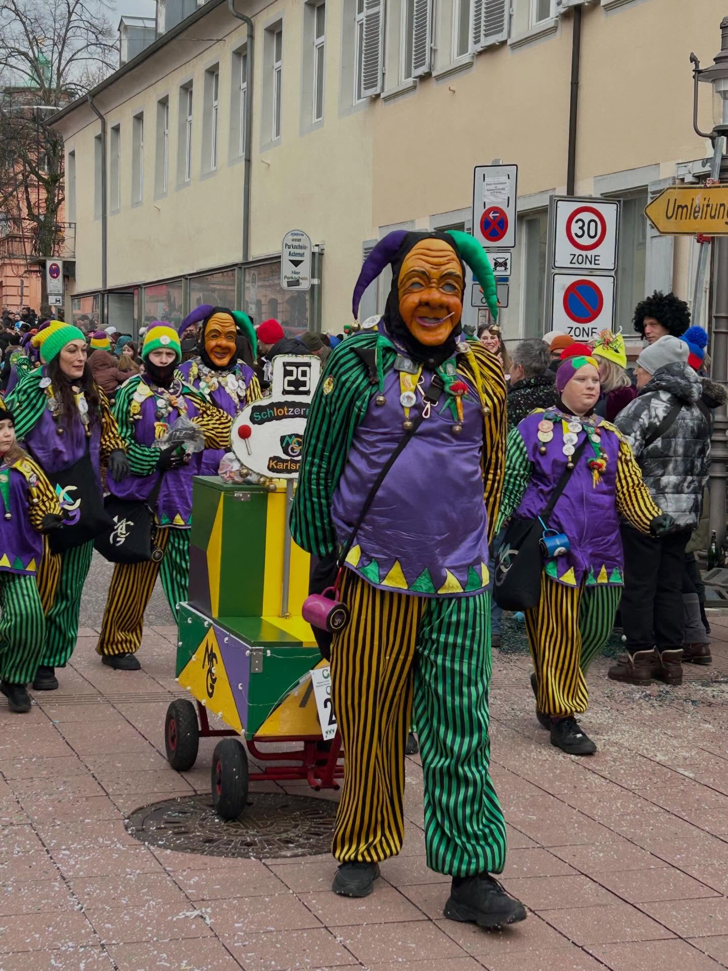 Dieser Alternativtext wurde KI-generiert: Eine Gruppe von Personen in farbenfrohen Kostümen zieht auf einer Straße. Im Hintergrund sind Gebäude und Verkehrsschilder zu sehen.