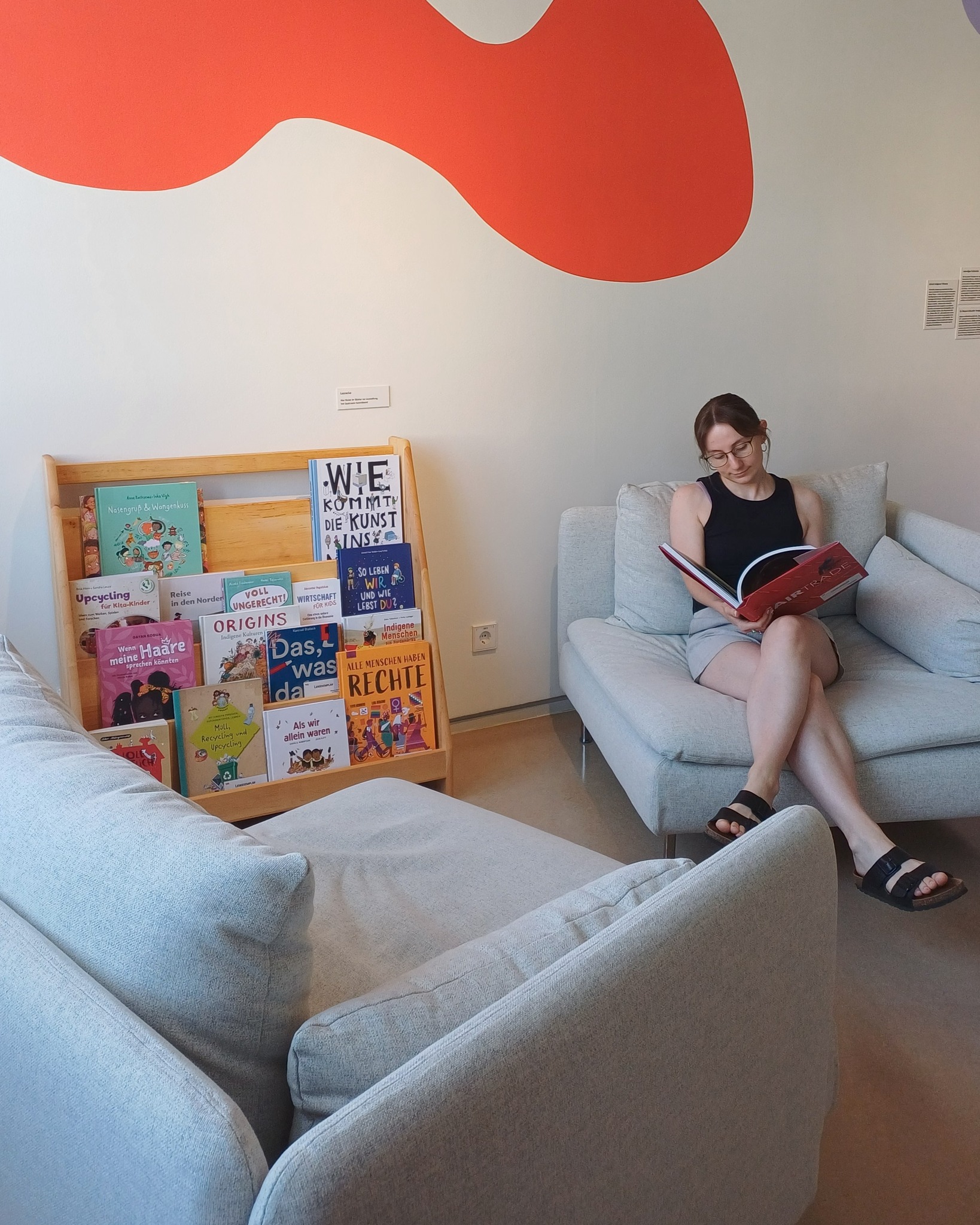 Eine Person sitzt auf einem hellgrauen Sofa und liest ein Buch mit rotem Einband. Neben ihr steht ein Holzregal mit Kinderbüchern, darunter Titel wie „Wie kommt die Kunst ins Museum?“ und „Alle Menschen haben Rechte“. An der Wand darüber ist ein großes, orangefarbenes abstraktes Formelement angebracht. (KI-Alternativtext)
