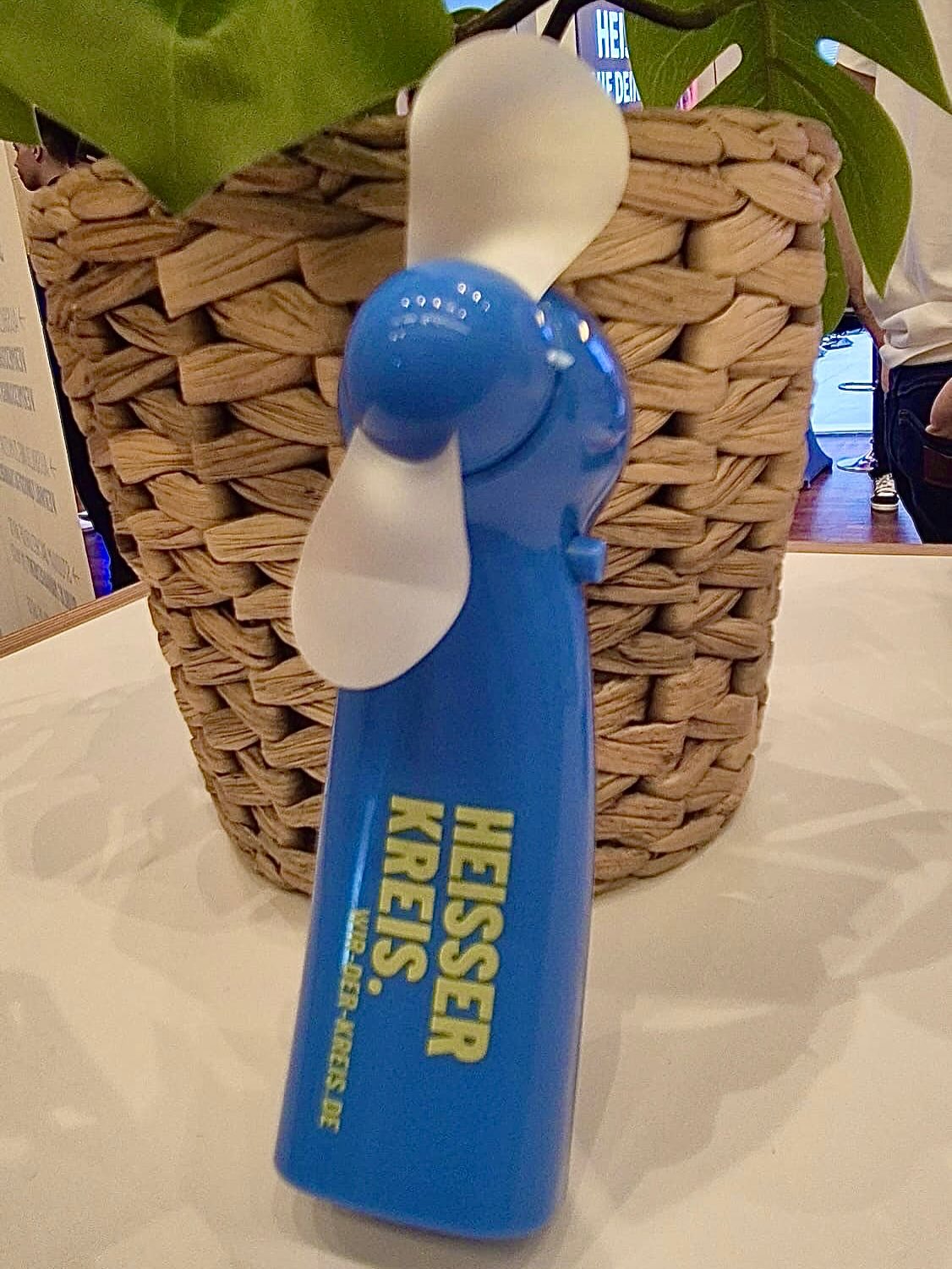 Ein blauer Handventilator mit weißen Flügeln steht vor einem geflochtenen Korb. Auf dem Griff ist in gelber Schrift der Text "HEISSER KREIS." sowie die Webadresse "WIR-DER-KREIS.DE" zu lesen. (KI-Alternativtext)