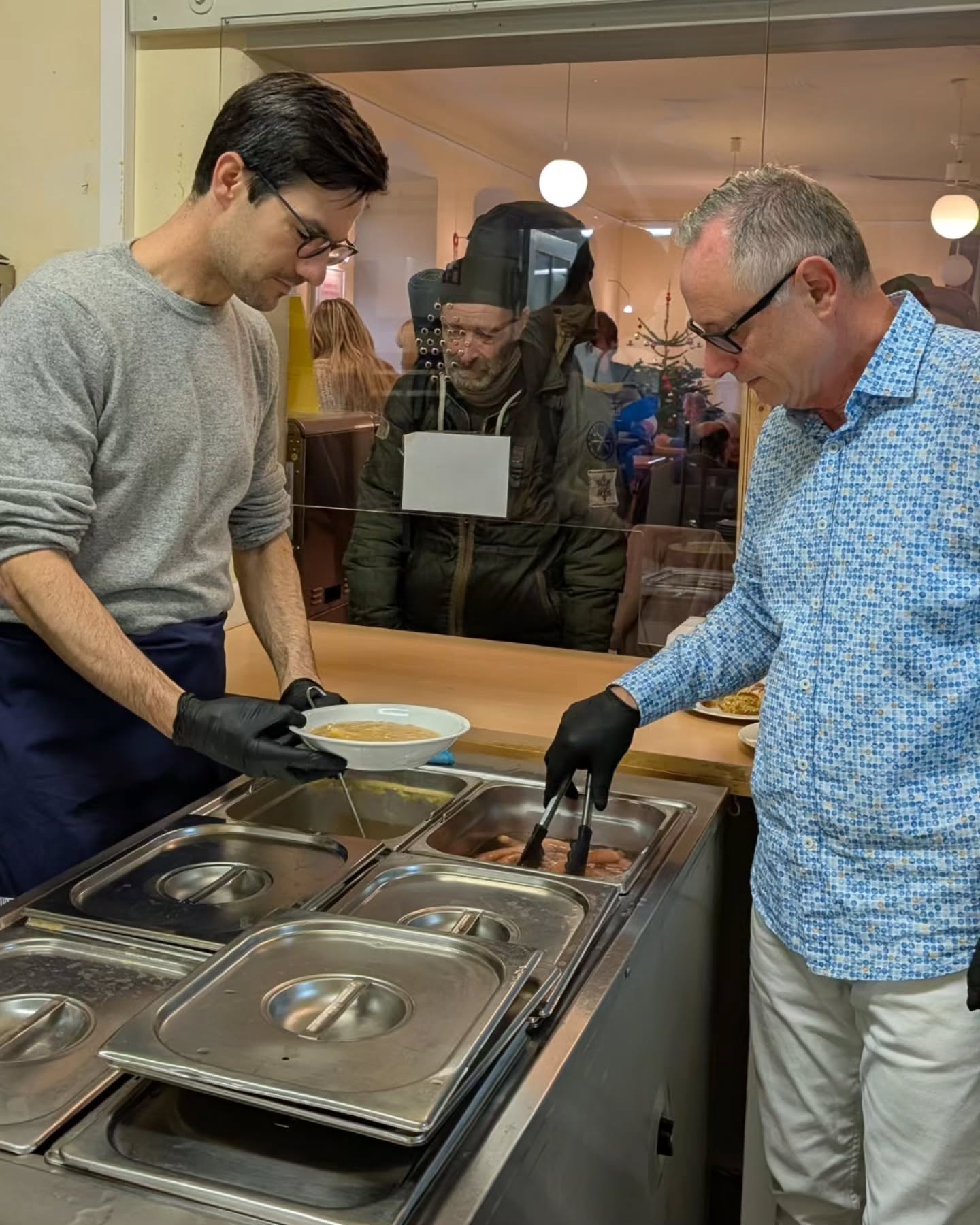 Dieser Alternativtext wurde KI-generiert: Zwei Personen servieren Essen an einem Buffet aus. Im Hintergrund sind weitere Personen in einem Innenraum zu sehen.