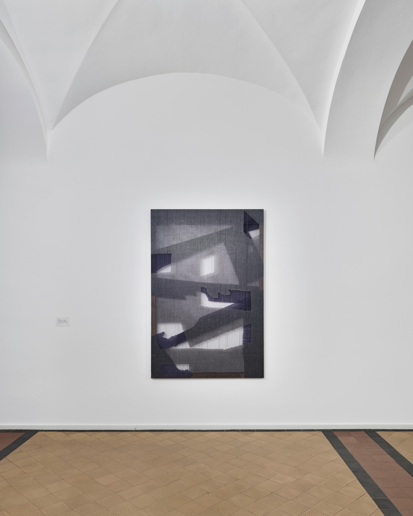 Dieser Alternativtext wurde KI-generiert: Ein abstraktes Gemälde in Grautönen hängt an einer hellen Wand. Der Raum hat einen gefliesten Boden und eine gewölbte Decke.