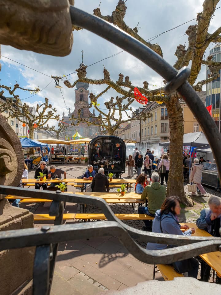 Dieser Alternativtext wurde KI-generiert: Eine Stadtansicht mit einem belebten Platz im Vordergrund. Im Hintergrund ist ein Kirchturm und Gebäude zu sehen, teilweise durch einen schmiedeeisernen Rahmen gerahmt.