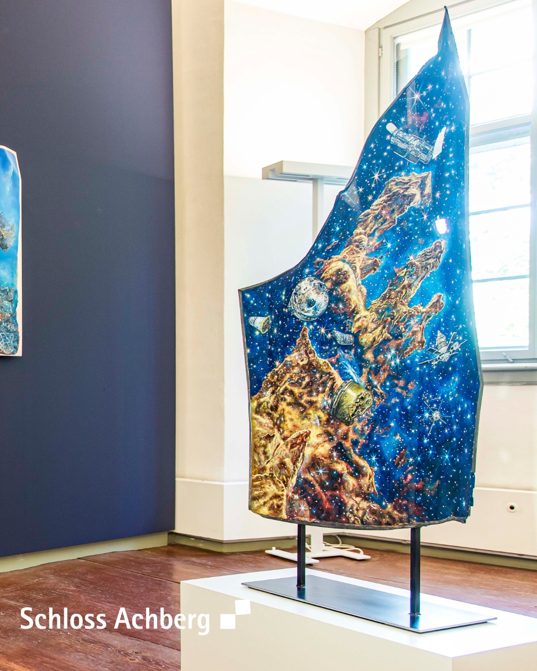 Dieser Alternativtext wurde KI-generiert: Ein farbenfrohes, vertikal angebrachtes Textil mit Weltraum-Motiven steht auf einem Metallgestell in einem Raum. Im Hintergrund ist ein Fenster und eine dunkel gestrichene Wand zu sehen.