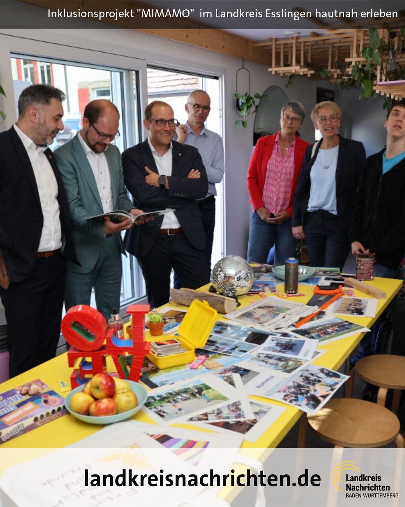 Eine Gruppe von Menschen steht um einen Tisch mit Gegenständen wie Spielzeug und Fotos. Auf dem Tisch steht der Schriftzug landkreisnachrichten.de.