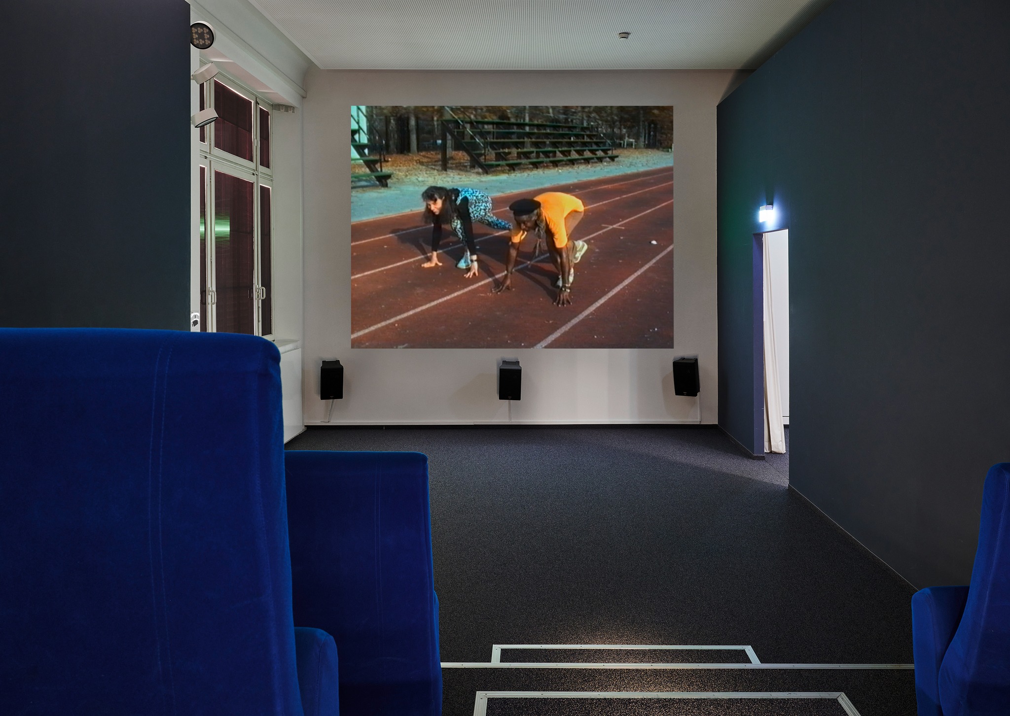 Ansicht eines kleinen Kinosaals mit blauen Sesseln im Vordergrund und dunklen Wänden. Auf der Leinwand sind zwei Personen in der Startposition auf einer Laufbahn zu sehen. Darunter sind Lautsprecher an der Wand angebracht. (KI-Alternativtext)