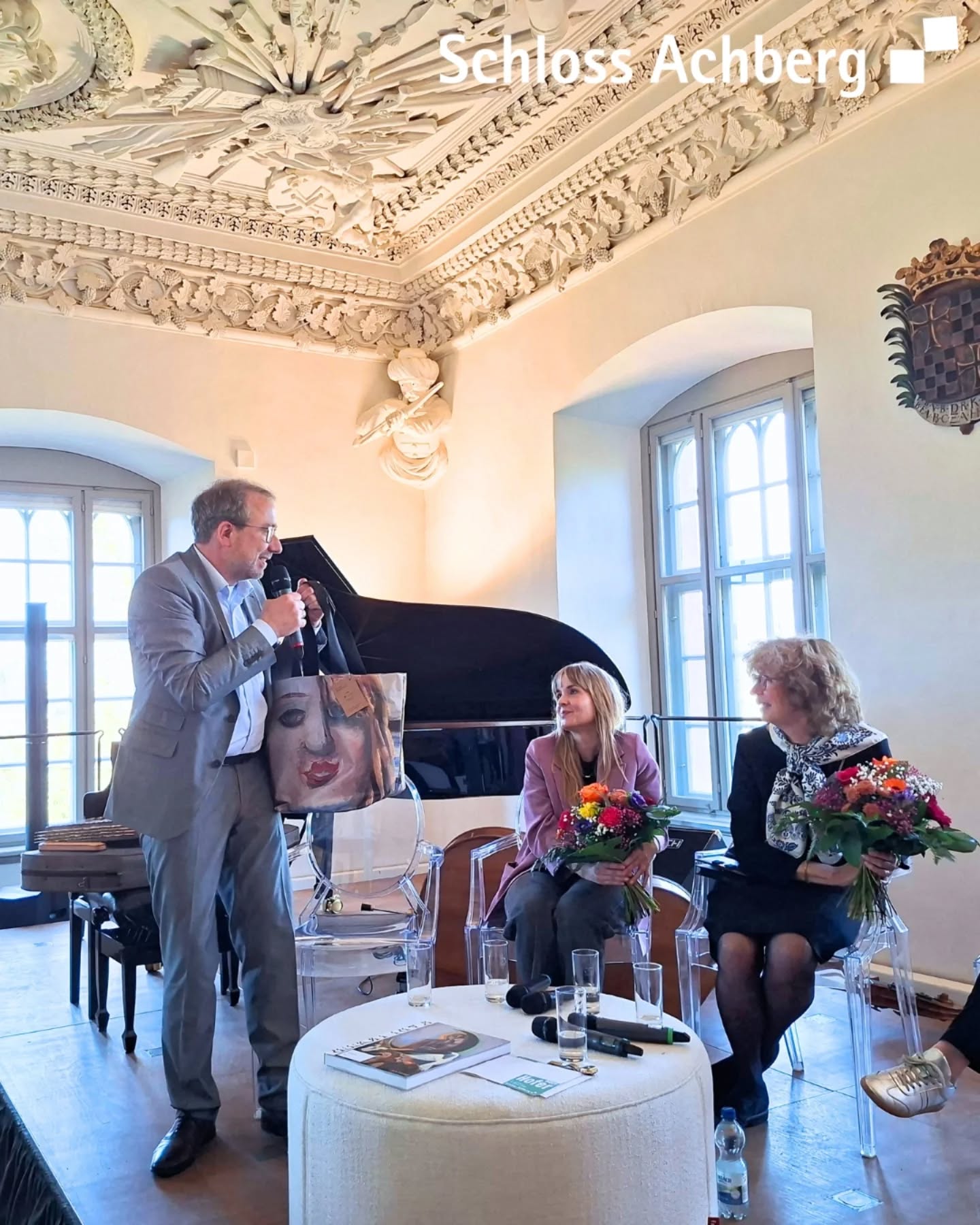 Dieser Alternativtext wurde KI-generiert: Ein Innenraum mit einer Person, die spricht, und anderen, die zuhören. Ein Klavier und Blumenarrangements sind ebenfalls im Bild zu sehen.