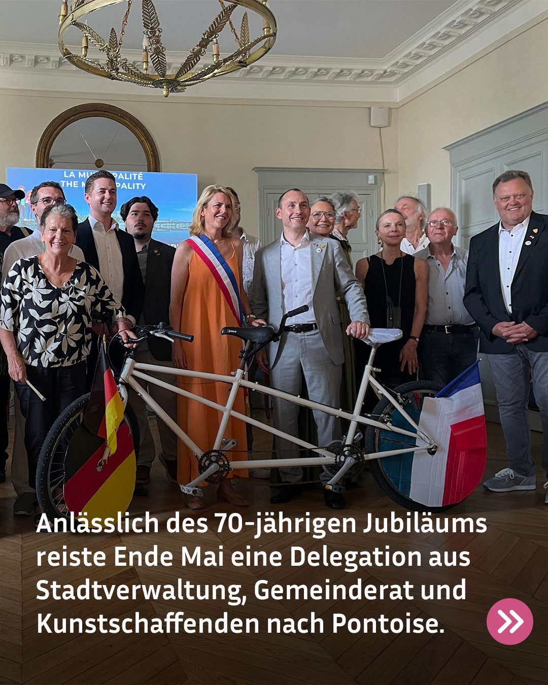 Eine Gruppe von Personen steht in einem Saal hinter einem langen weißen Tandemrad, an dem vorne die deutsche und hinten die französische Flagge befestigt ist. Eine Person in der Mitte trägt eine orange Amtsschärpe. Ein Textfeld unten lautet: „Anlässlich des 70-jährigen Jubiläums reiste Ende Mai eine Delegation aus Stadtverwaltung, Gemeinderat und Kunstschaffenden nach Pontoise.“ (KI-Alternativtext)