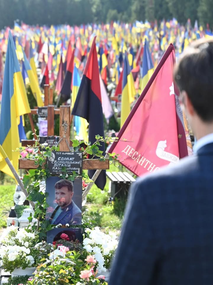 Dieser Alternativtext wurde KI-generiert: Ein Grab mit Blumen und Fahnen im Freien. Im Hintergrund ist eine Person zu sehen, die dem Grab zugewandt ist.