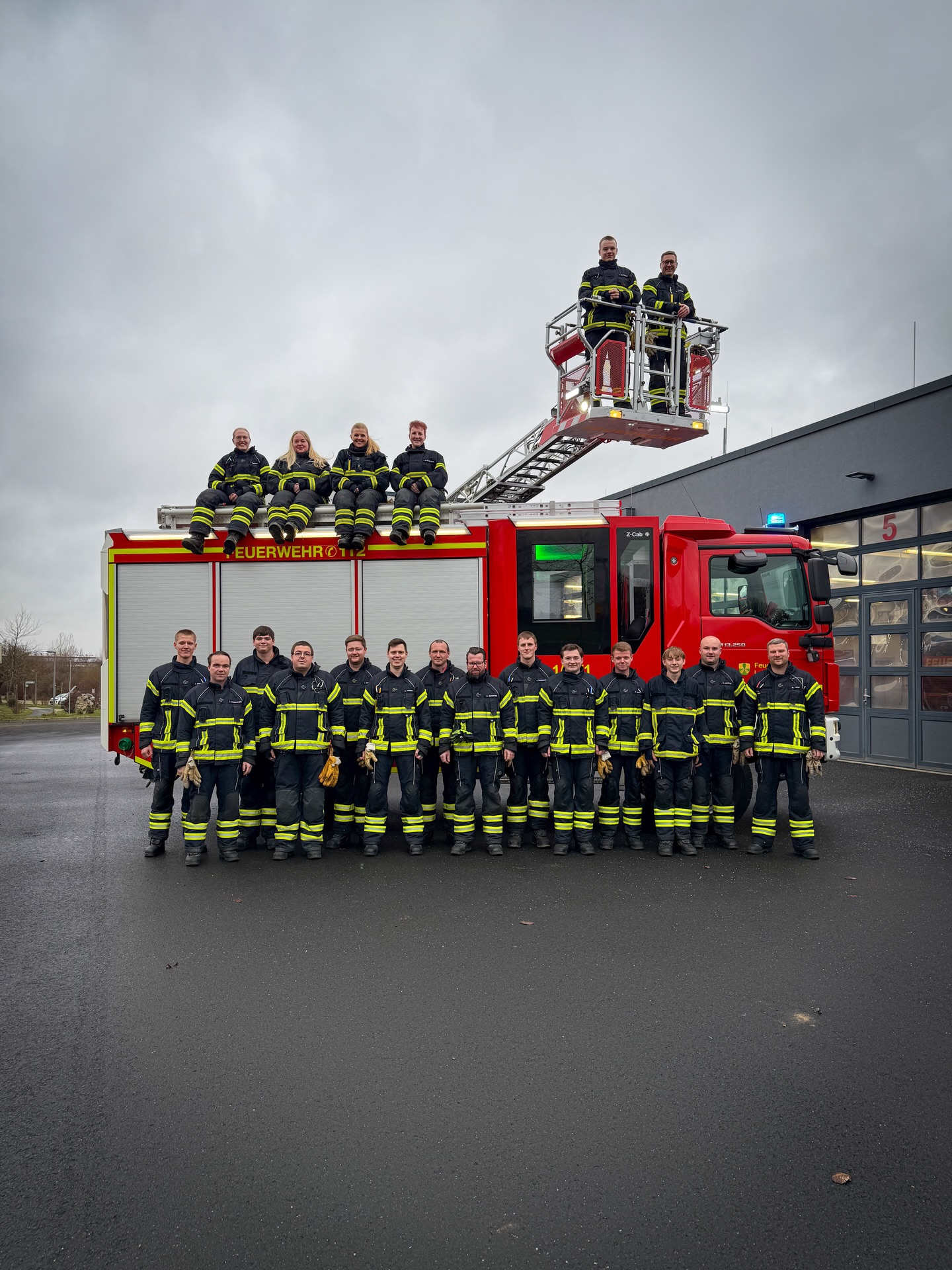 Eine Gruppe von Personen in Feuerwehruniform posiert vor einem roten Einsatzfahrzeug mit ausgefahrener Leiter. Vier Personen sitzen auf dem Fahrzeugaufbau, zwei stehen im Korb der Leiter. Auf der Seite des Fahrzeugs ist der Schriftzug "FEUERWEHR OTTZ" zu sehen. (KI-Alternativtext)