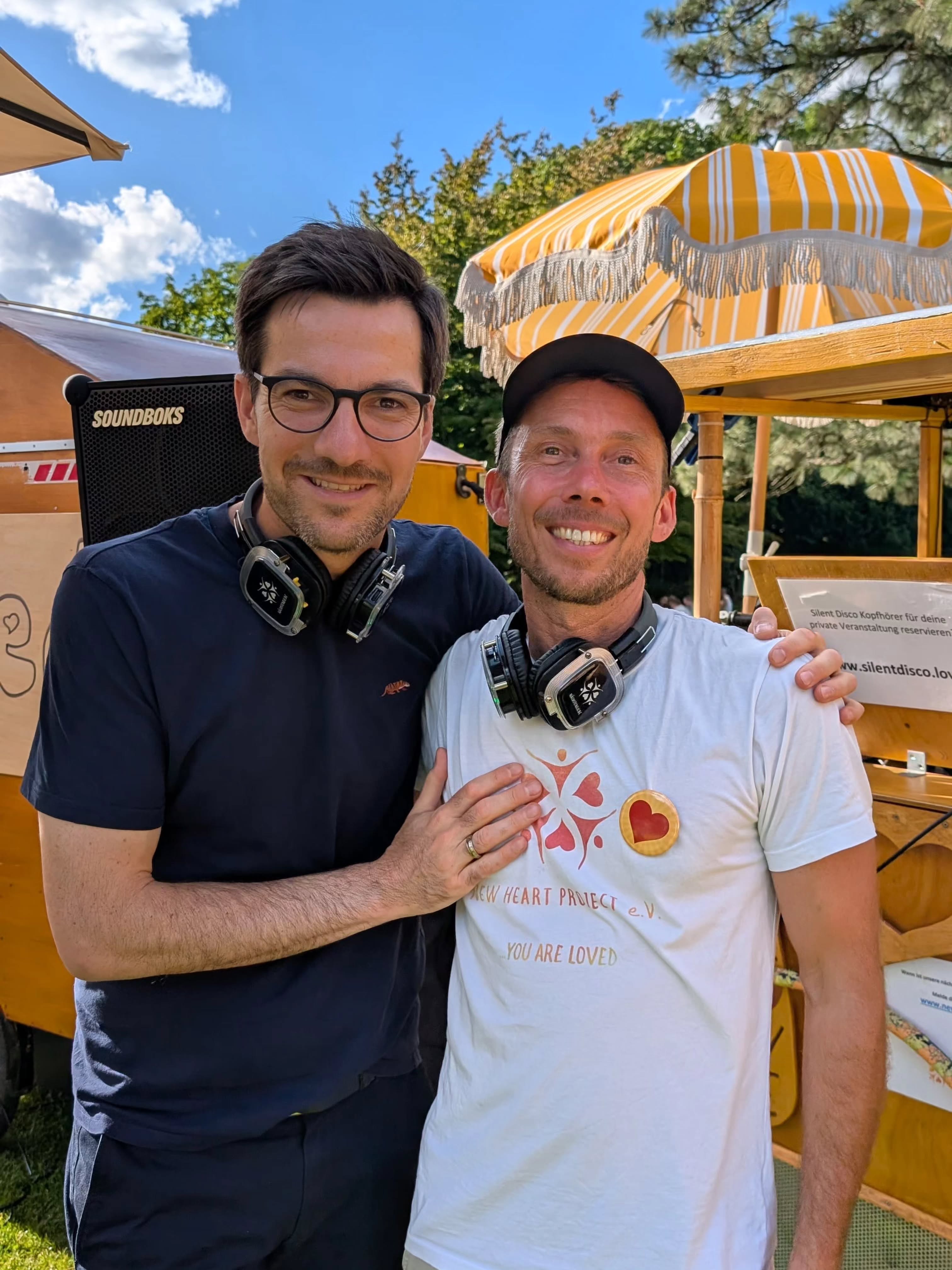 Zwei lächelnde Personen mit Kopfhörern um den Hals stehen im Freien vor einem Stand mit gelb-weiß gestreifter Markise. Eine trägt ein weißes T-Shirt mit dem Aufdruck „NEW HEART PROJECT e.V. ...YOU ARE LOVED“ und einem Herz-Abzeichen, die andere ein dunkles Shirt. Im Hintergrund ist ein Lautsprecher mit der Aufschrift „SOUNDBOKS“ und ein Schild mit „Silent Disco Kopfhörer für deine private Veranstaltung reservieren“ zu sehen. (KI-Alternativtext)