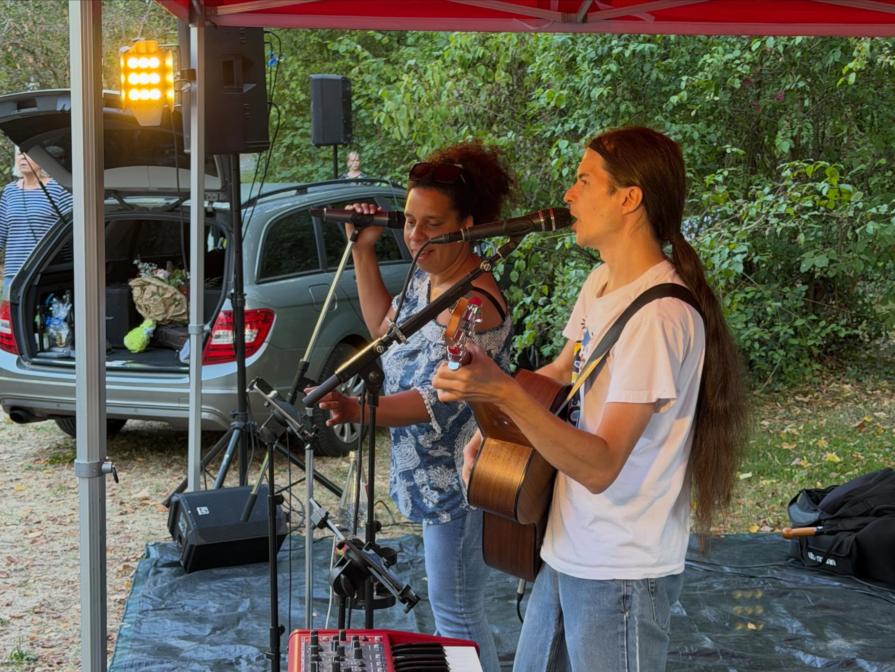 Zwei Personen spielen unter einem roten Zeltdach Musik. Eine Person singt in ein Mikrofon, die andere spielt eine Akustikgitarre und singt ebenfalls. Im Vordergrund steht ein rotes Keyboard, im Hintergrund ein Auto mit offenem Kofferraum. (KI-Alternativtext)