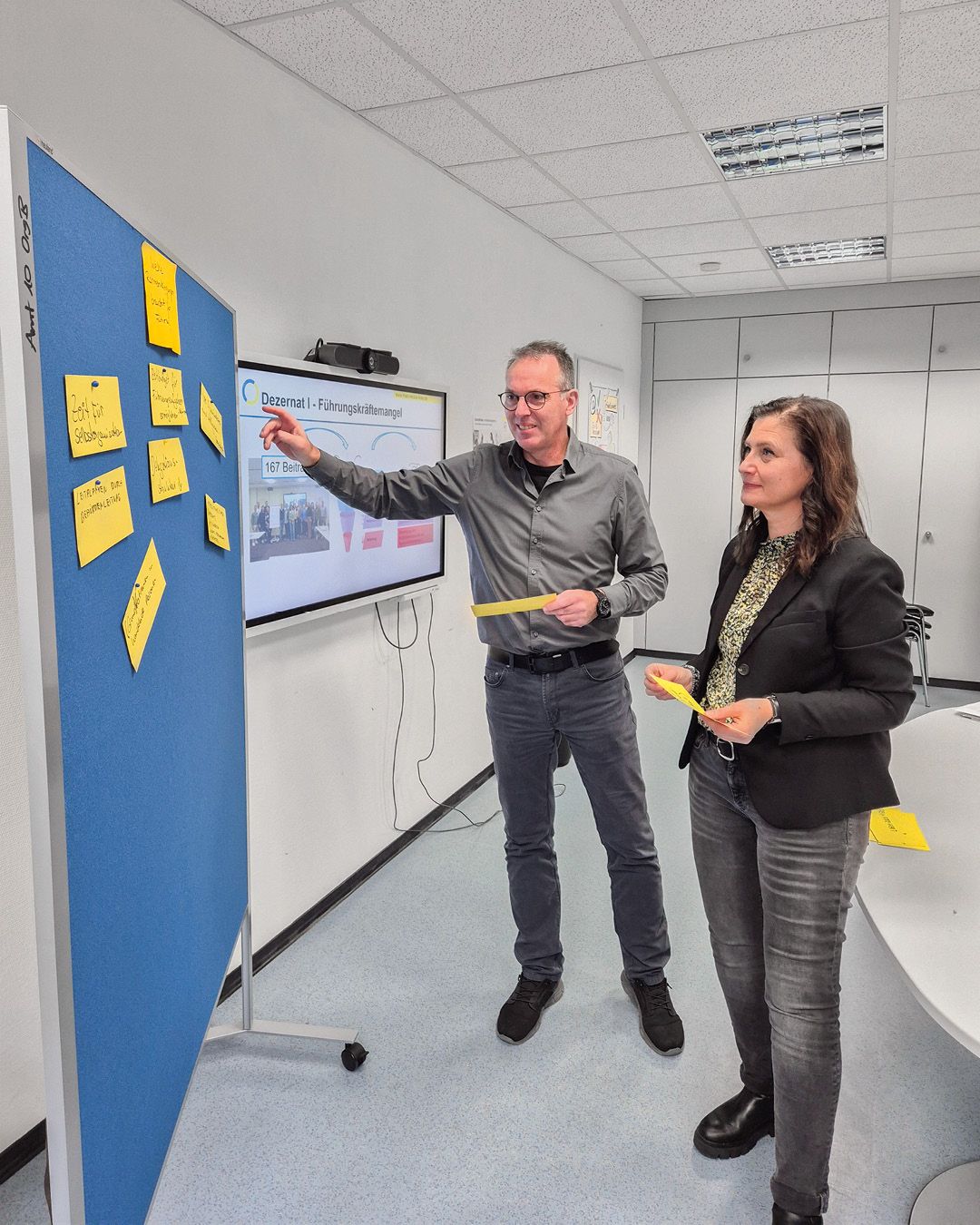 Dieser Alternativtext wurde KI-generiert: Zwei Personen stehen in einem Büro vor einer blauen Pinnwand mit Haftnotizen und einem Diagramm. Eine Person zeigt mit dem Finger auf das Diagramm.