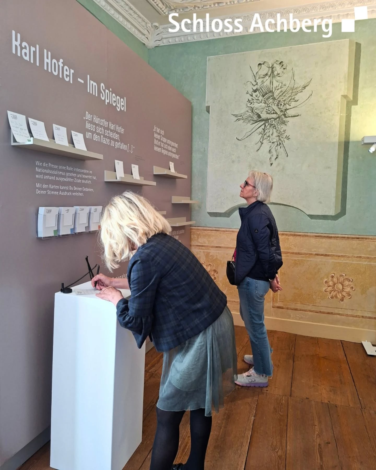 Dieser Alternativtext wurde KI-generiert: Ein Raum mit einer Ausstellung an der Wand. Eine Person steht mit dem Rücken zum Betrachter vor der Ausstellung und betrachtet diese.