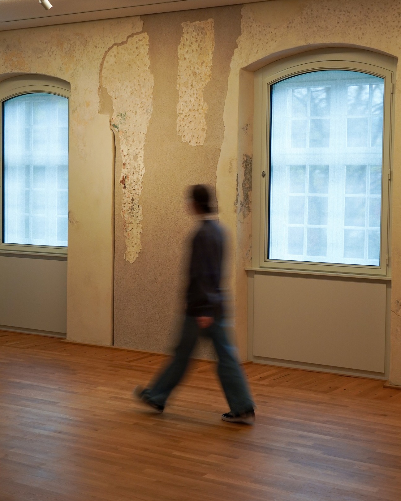 Dieser Alternativtext wurde KI-generiert: Ein Innenraum mit einer Wand, an der Putz abblättert. Eine Person geht verschwommen vorbei, während Tageslicht durch Fenster scheint.