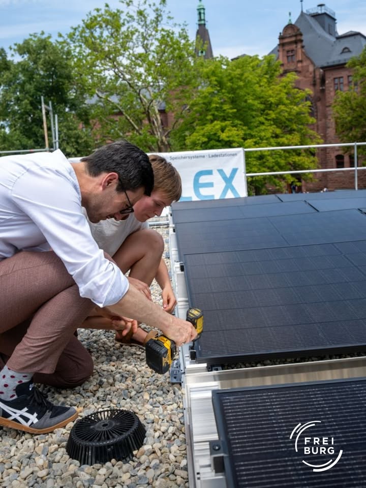 Zwei Personen knien auf einem Kiesboden und montieren Solarpanels auf einem Metallgestell. Eine Person verwendet einen gelben Akkuschrauber, während die andere assistiert. Im Hintergrund ist ein weißes Banner mit der Aufschrift „LEX“ und „Speichersysteme · Ladestationen“ vor einem historischen Backsteingebäude zu sehen. (KI-Alternativtext)