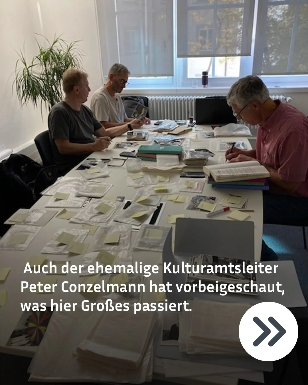 Drei Personen sitzen an einem langen Tisch in einem hellen Raum und bearbeiten Dokumente, Fotos in Plastiktüten mit gelben Post-its sowie eine offene Buchreihe. Eine Person rechts schreibt auf einer Stapel von Büchern. Im unteren Bildbereich steht der Text: "Auch der ehemalige Kulturamtsleiter Peter Conzelmann hat vorbeigeschaut, was hier Großes passiert." (KI-Alternativtext)