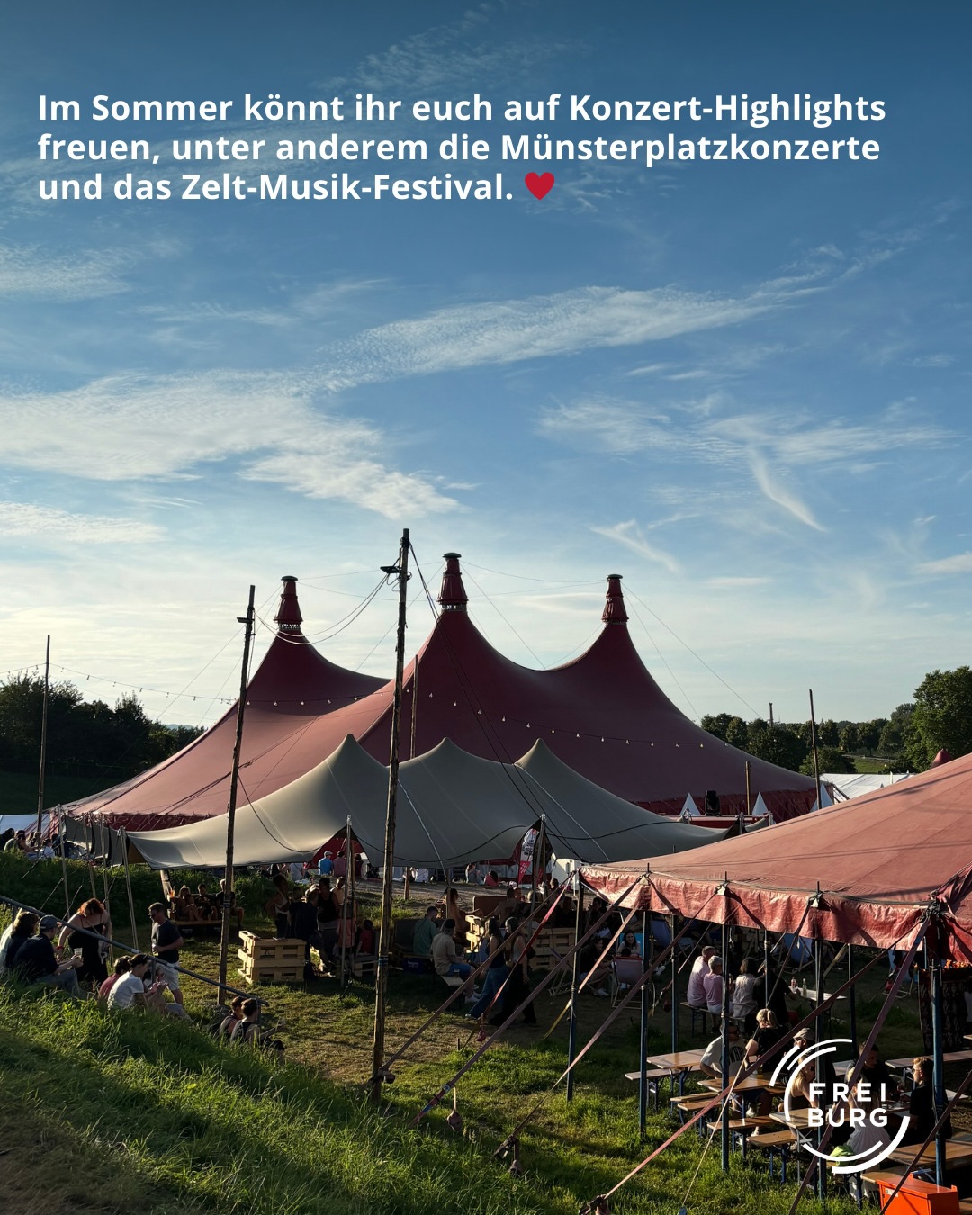 Dieser Alternativtext wurde KI-generiert: Ein Zelt steht auf einem Feld unter blauem Himmel. Im Hintergrund sind Menschen zu erkennen und im Vordergrund eine Anzeige für ein Festival.