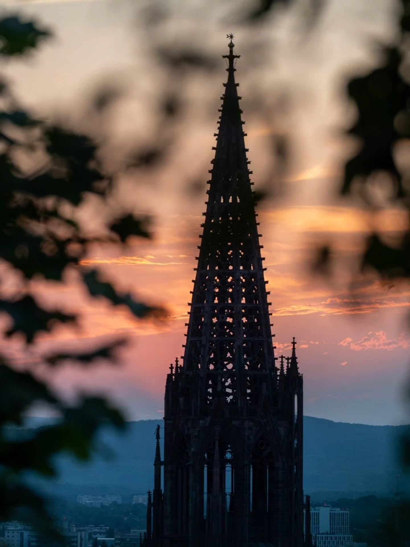 Ein hoher, gotischer Kirchturm steht in dunkler Silhouette vor einem orange-rosa gefärbten Abendhimmel. Unschärfe Blätter im Vordergrund rahmen die Szene ein, während im Hintergrund Hügel und Gebäude einer Stadt zu erkennen sind. (KI-Alternativtext)